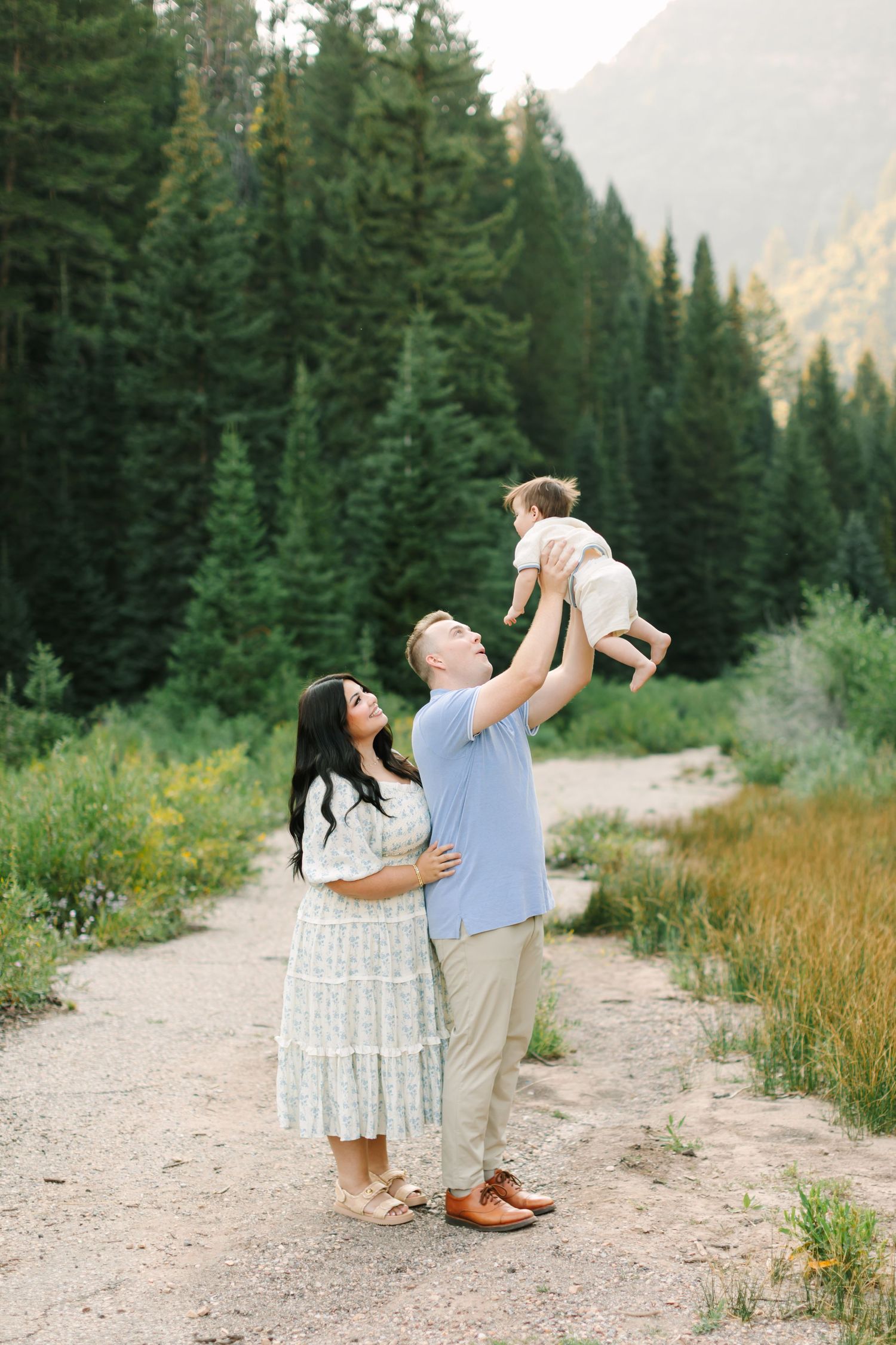 A family of three poses outdoors, dad lifting baby up while mom embraces him, surrounded by tall pine trees in Utah.