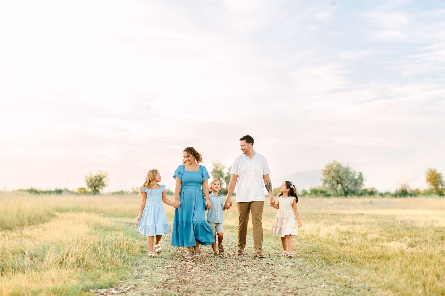Family of five walking hand-in-hand in a sunny field, wearing coordinated blue and white outfits in Bountiful, Utah.