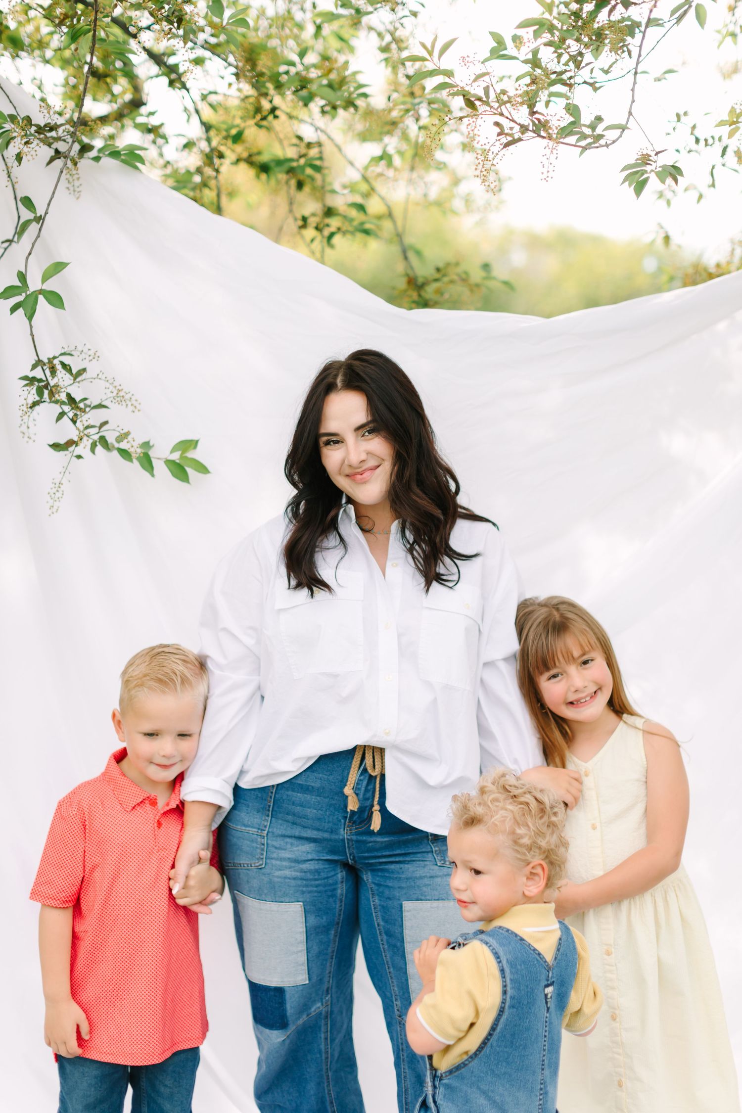 A woman in white shirt and jeans poses outdoors with three young children against a white fabric backdrop.