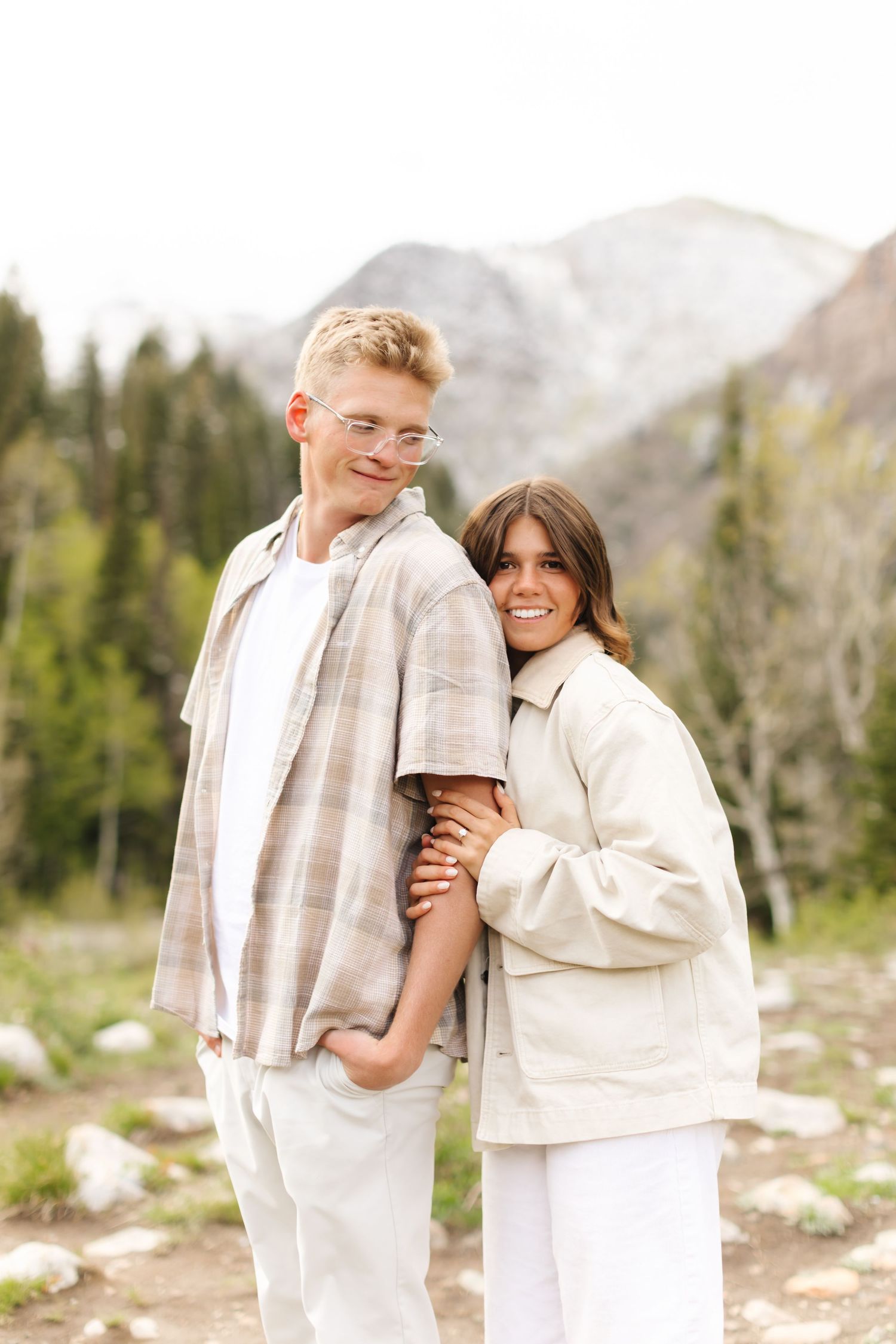 A young couple poses together outdoors with mountain and forest scenery, both wearing neutral tones, smiling warmly.
