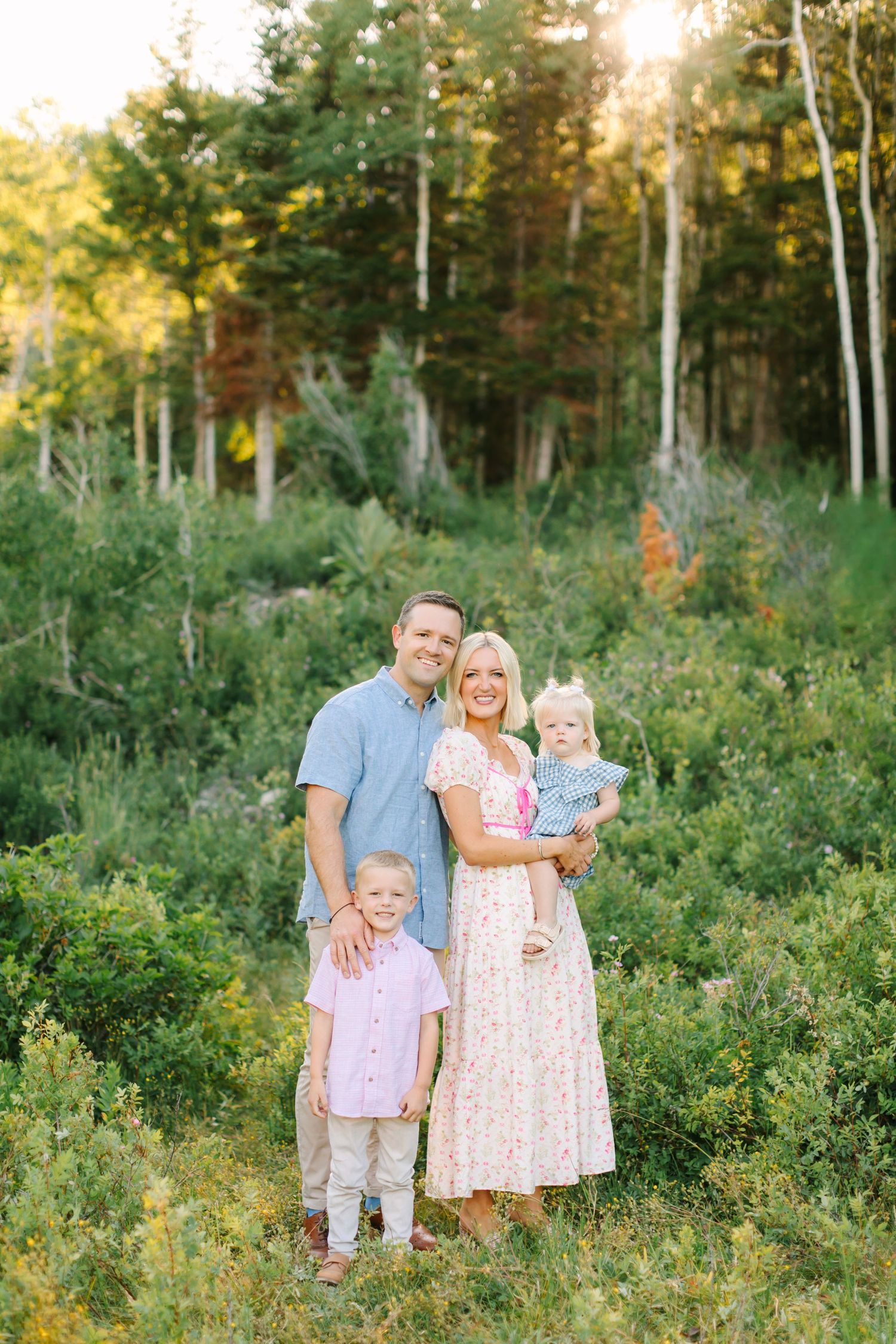Family of four poses in a sunlit forest clearing, parents smiling with two young children amid lush green foliage in American Fork Utah.