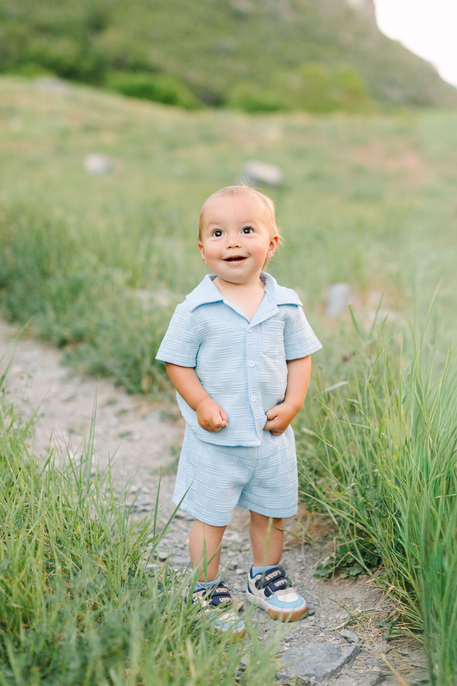 Smiling toddler boy in light blue shorts set standing on a dirt path surrounded by green grass and hills.
