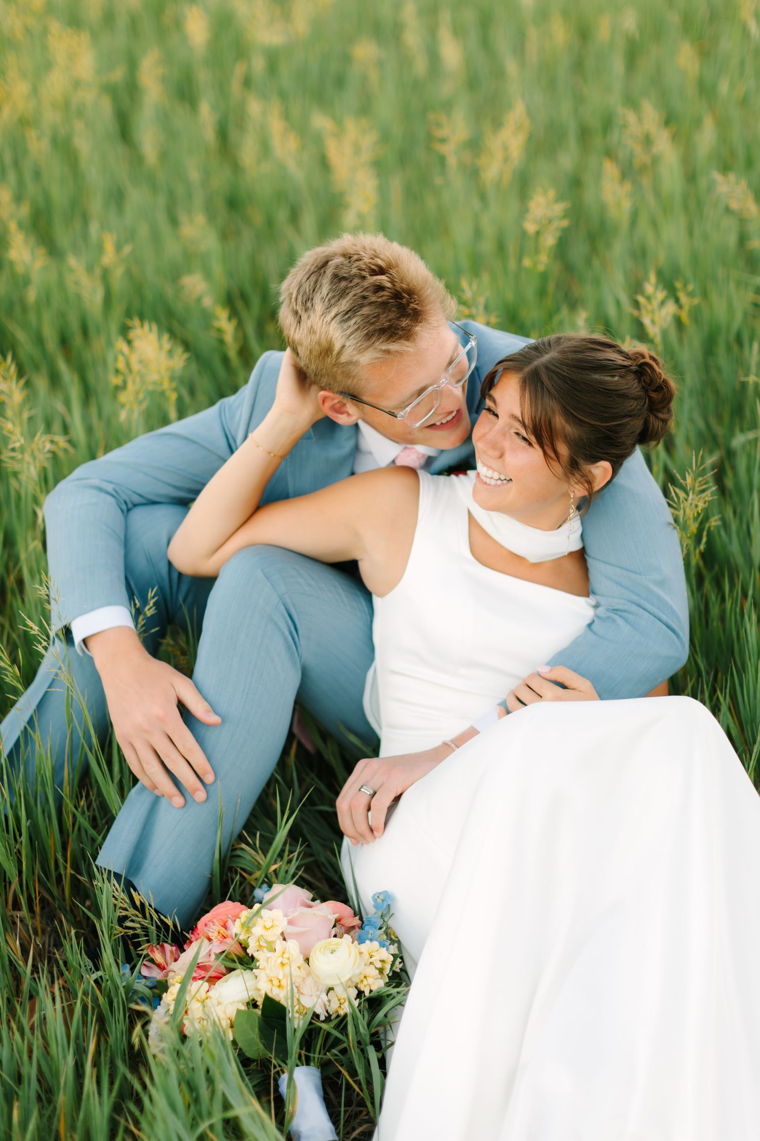 A smiling bride in white dress and groom in light blue suit sit together in a lush green field with a floral bouquet.