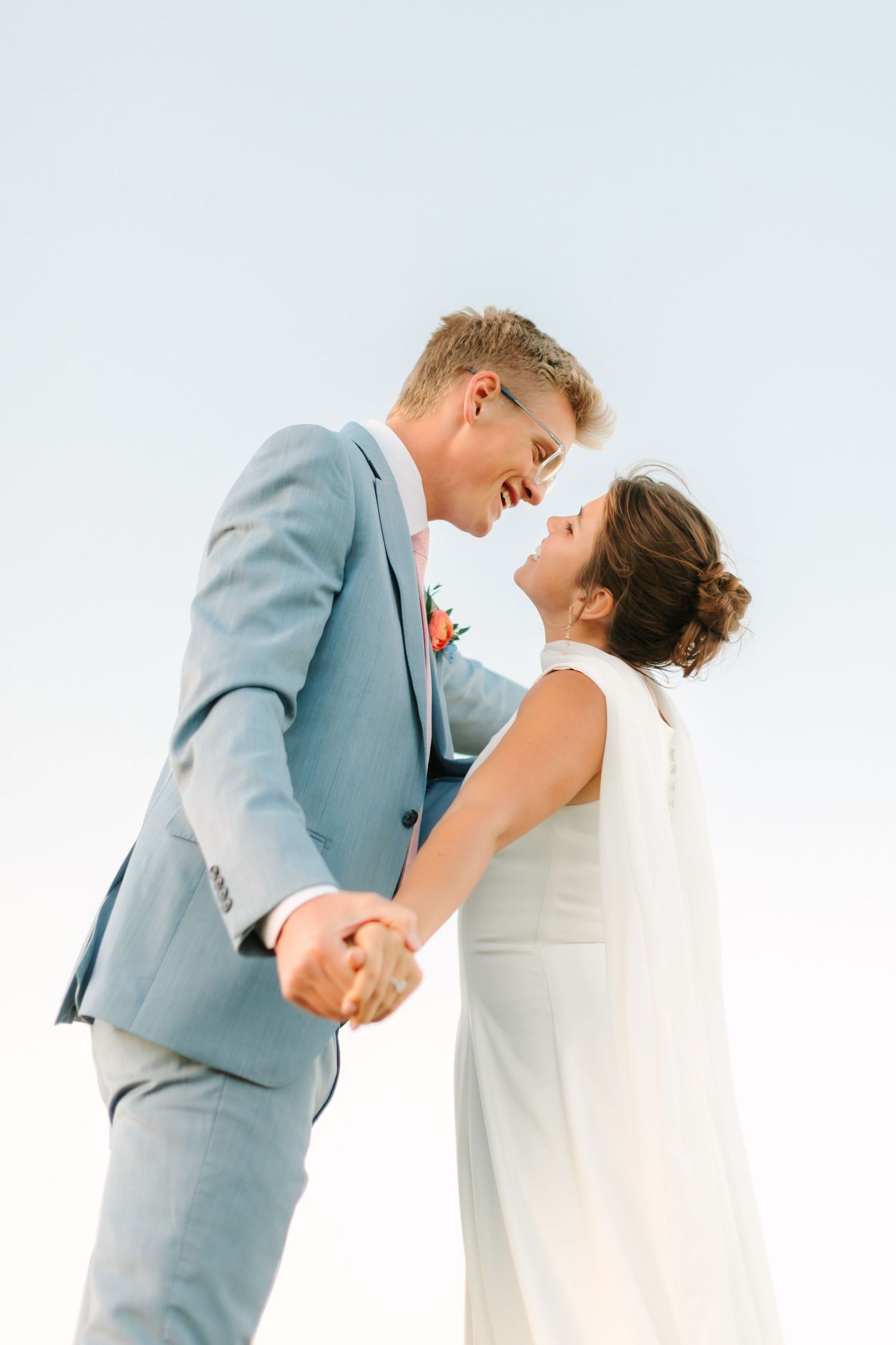 A joyful bride and groom in wedding attire hold hands and smile at each other against a clear blue sky.