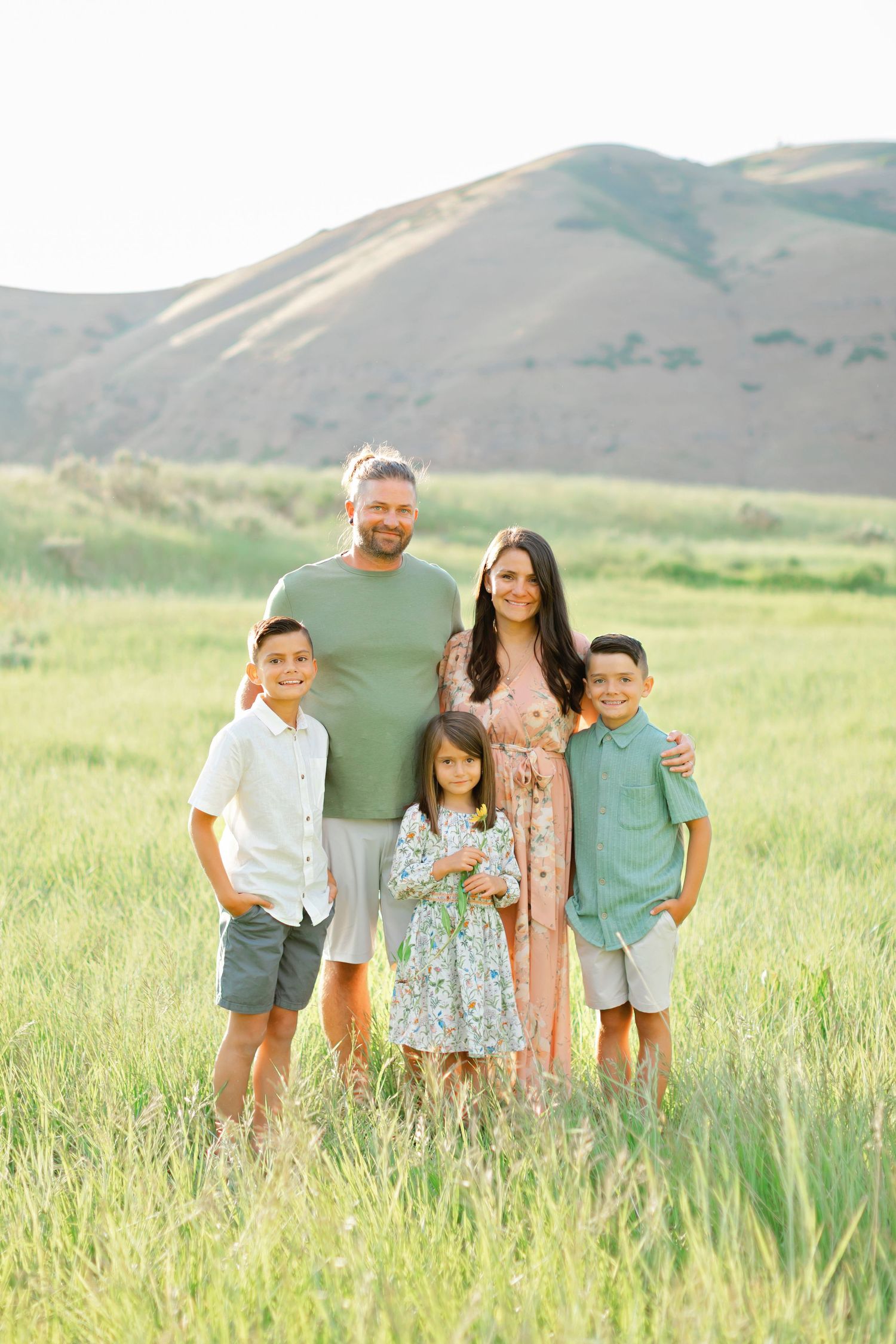 Family stands together in a sunlit meadow with rolling hills in the background during golden hour.