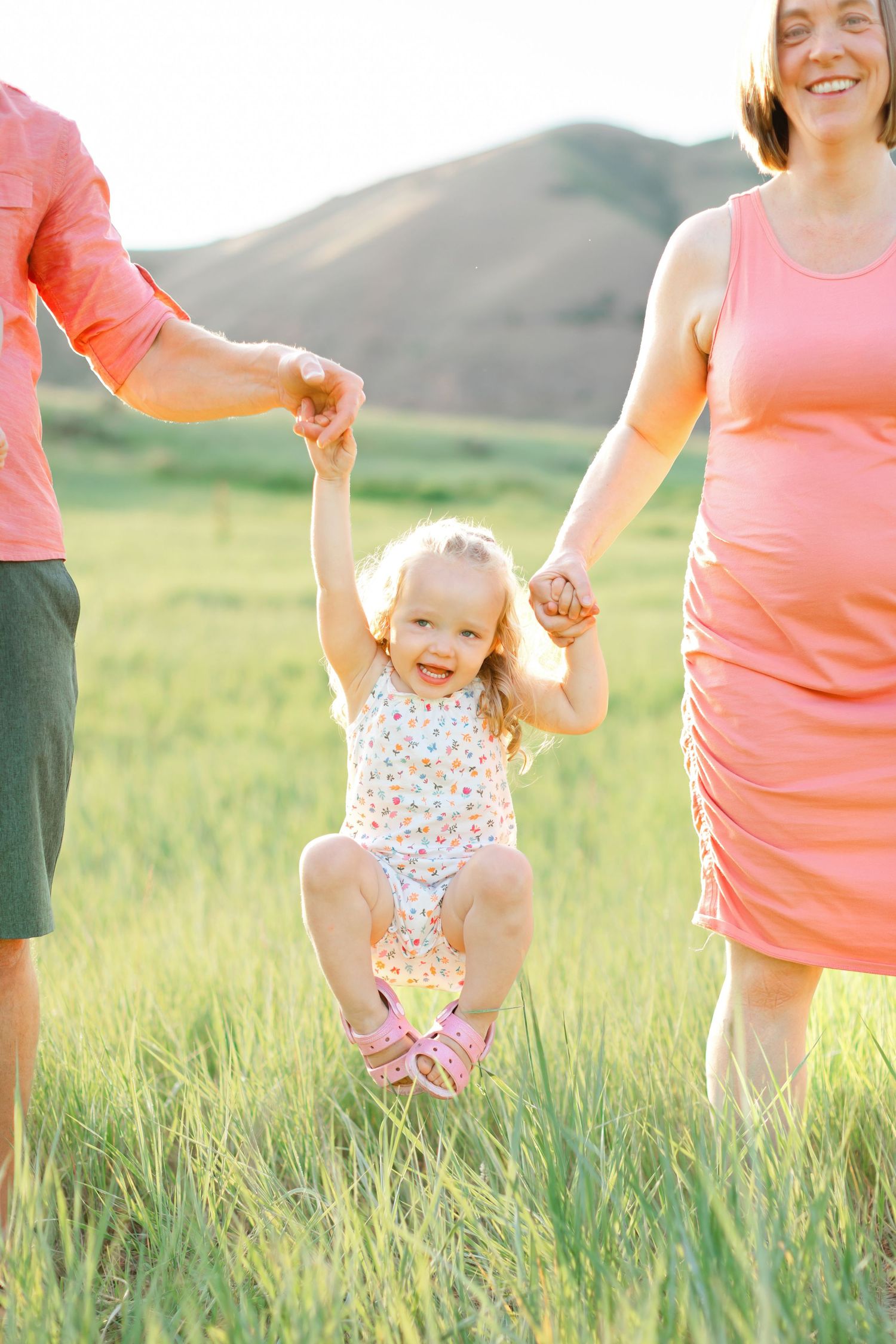A child in a floral swimsuit swings joyfully between two adults in pink clothing while standing in a grassy meadow.