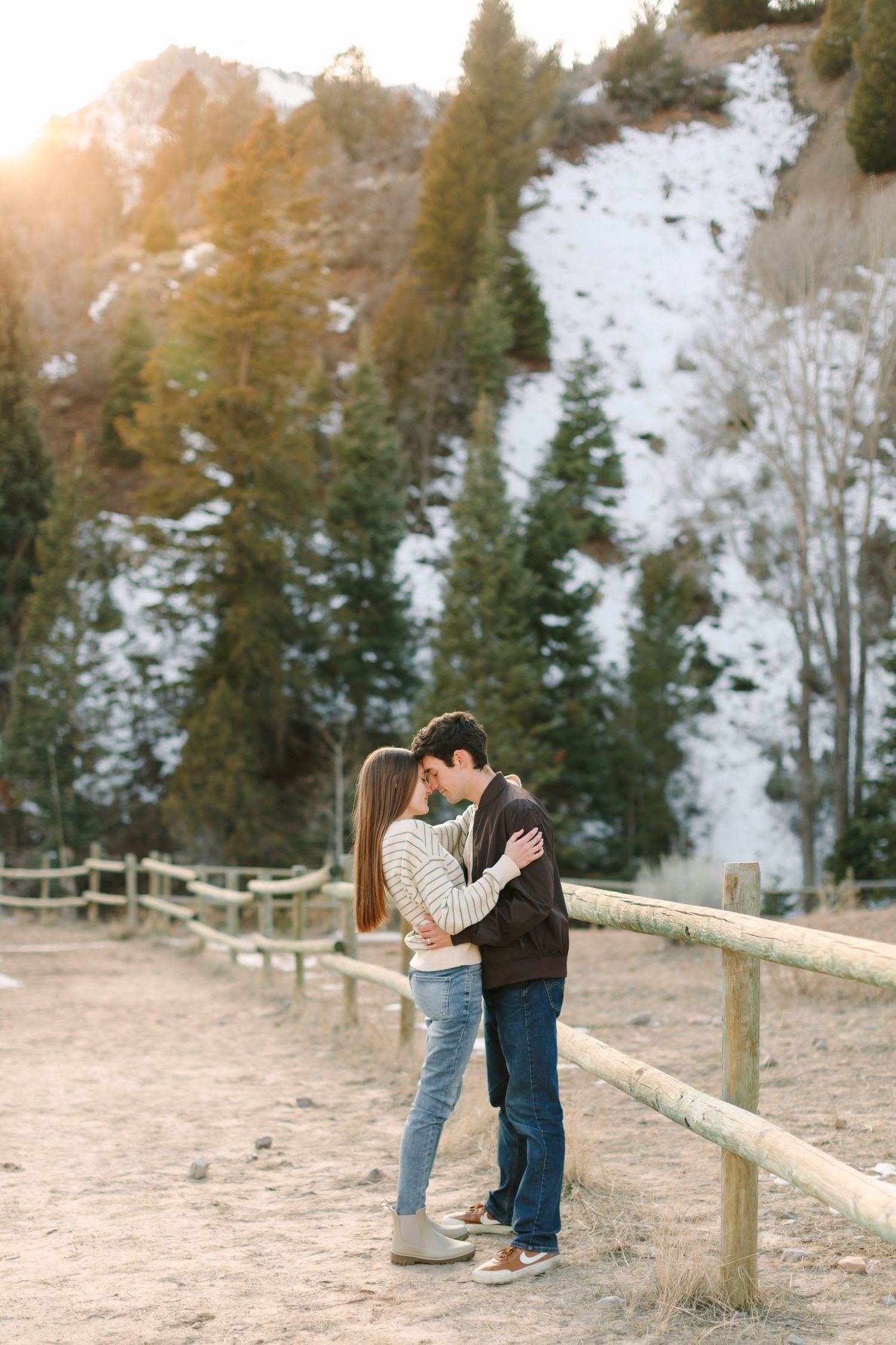 A couple shares a romantic moment together on a snowy winter path with evergreen trees in the background.