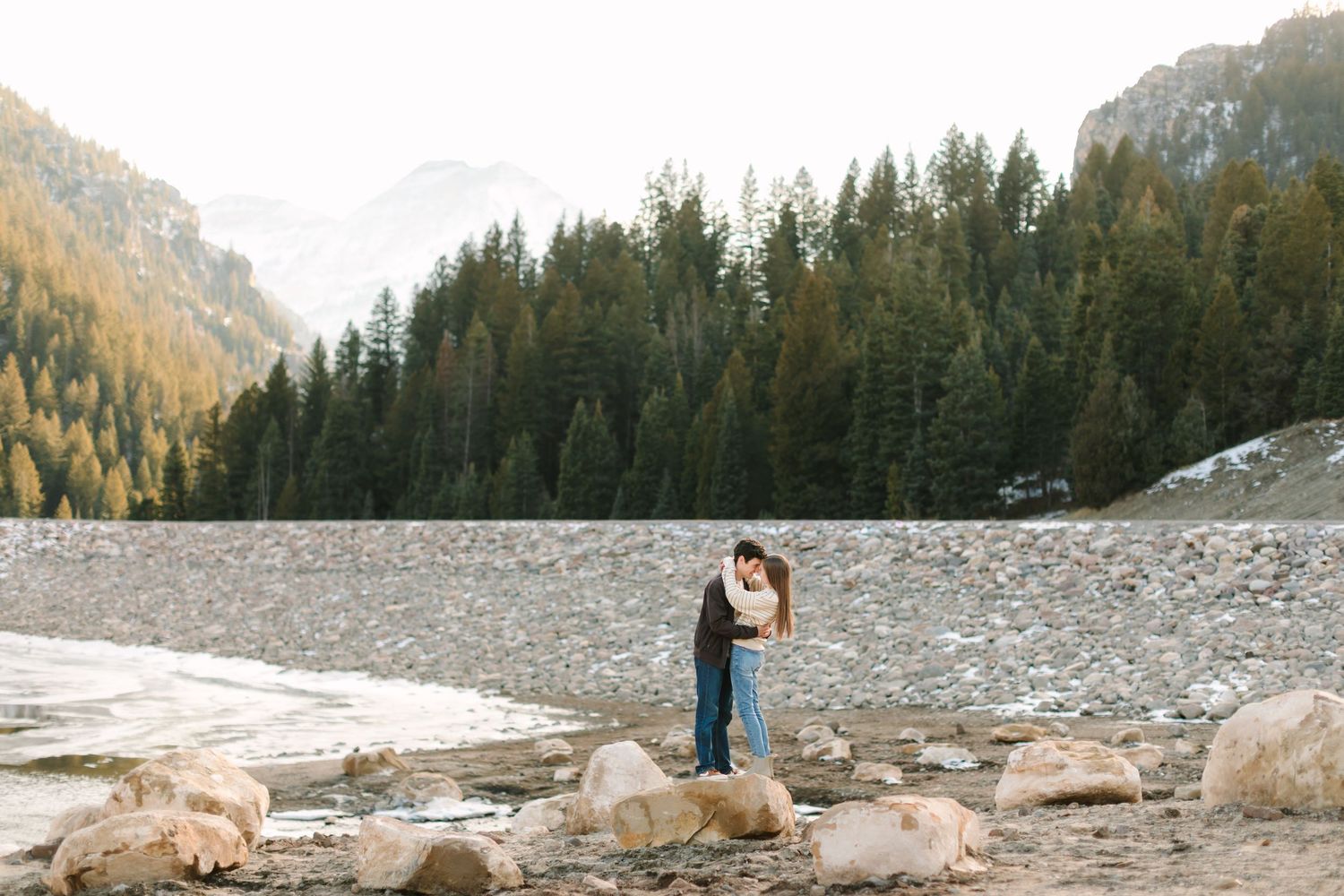 A couple embraces on rocky shoreline surrounded by evergreen forest and mountains in a scenic wilderness setting.