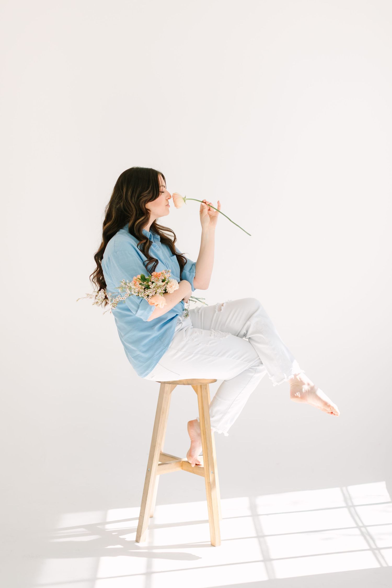 A creative portrait of someone in blue and white posing playfully on a wooden stool with natural light.