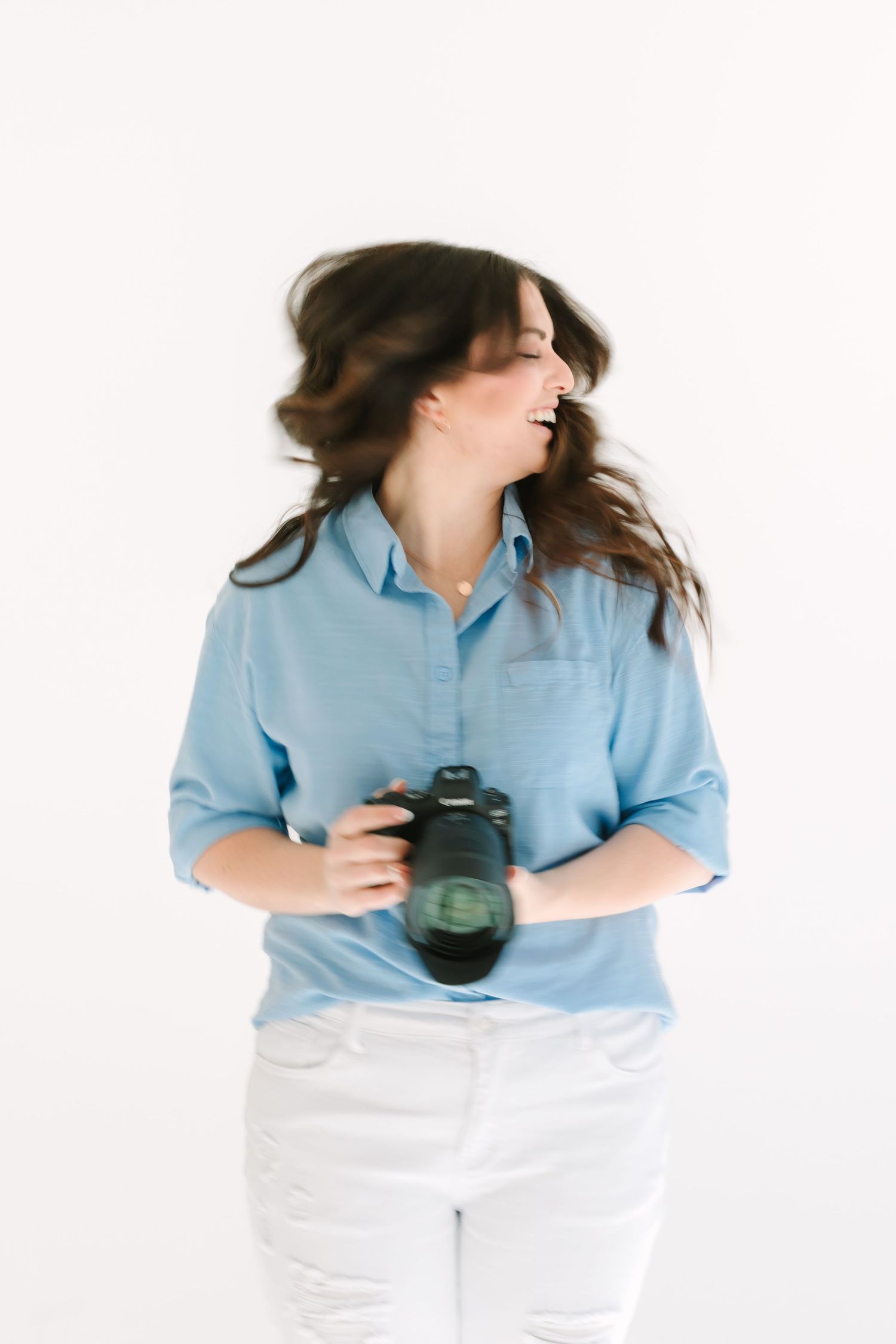 Photographer in light blue button-down shirt and white pants holds camera while hair flows in motion against white background.