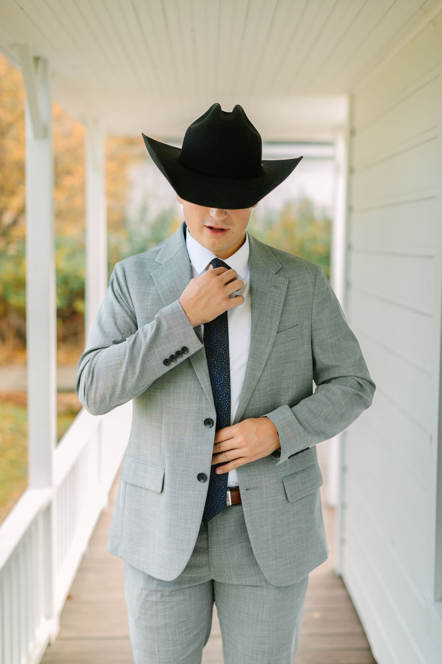 A figure in a light gray suit and black hat poses on a white porch in multiple stylish poses.