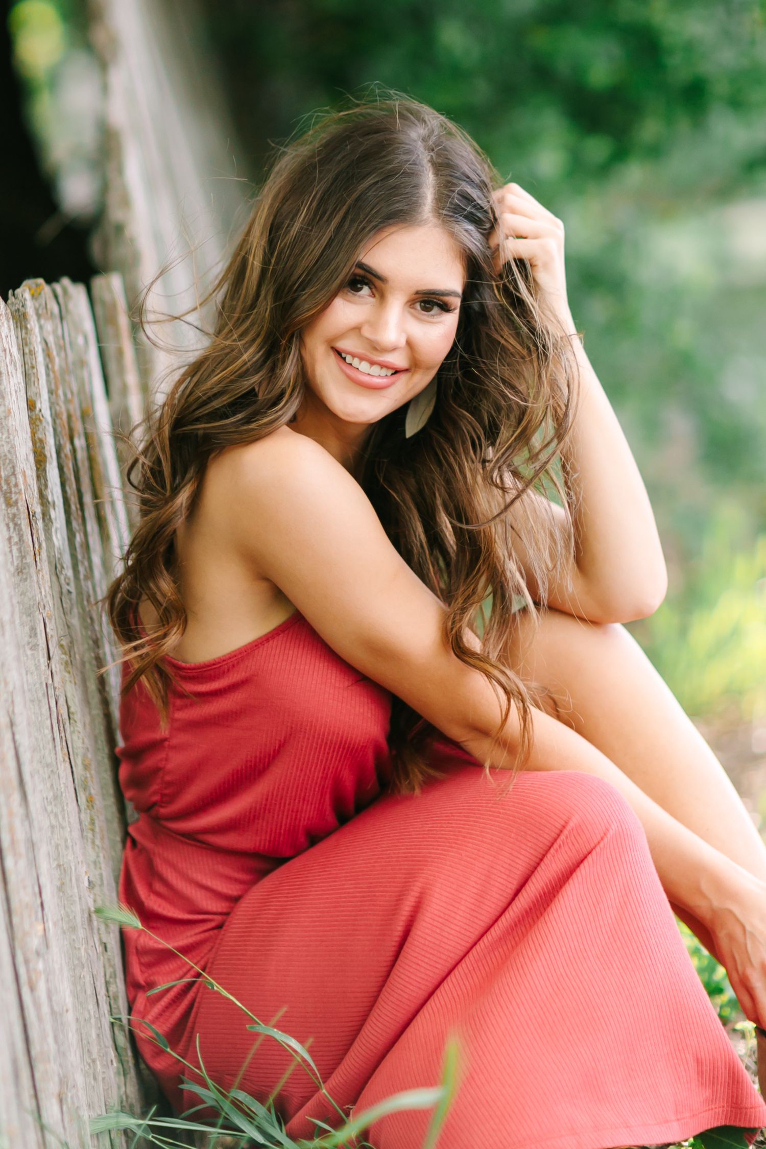 A woman in a coral dress sits outdoors against a rustic wooden fence in a natural setting.