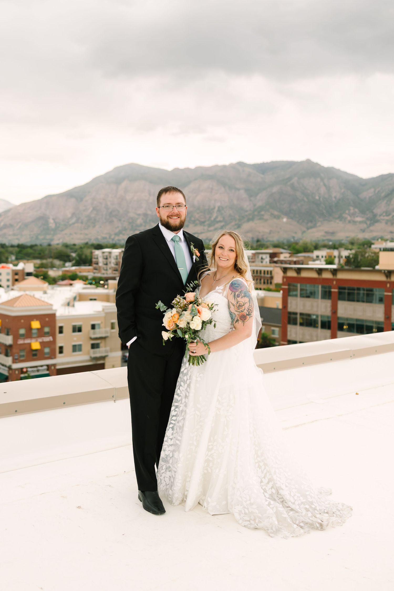 A bride and groom pose together on a rooftop with mountains and city buildings in the background.