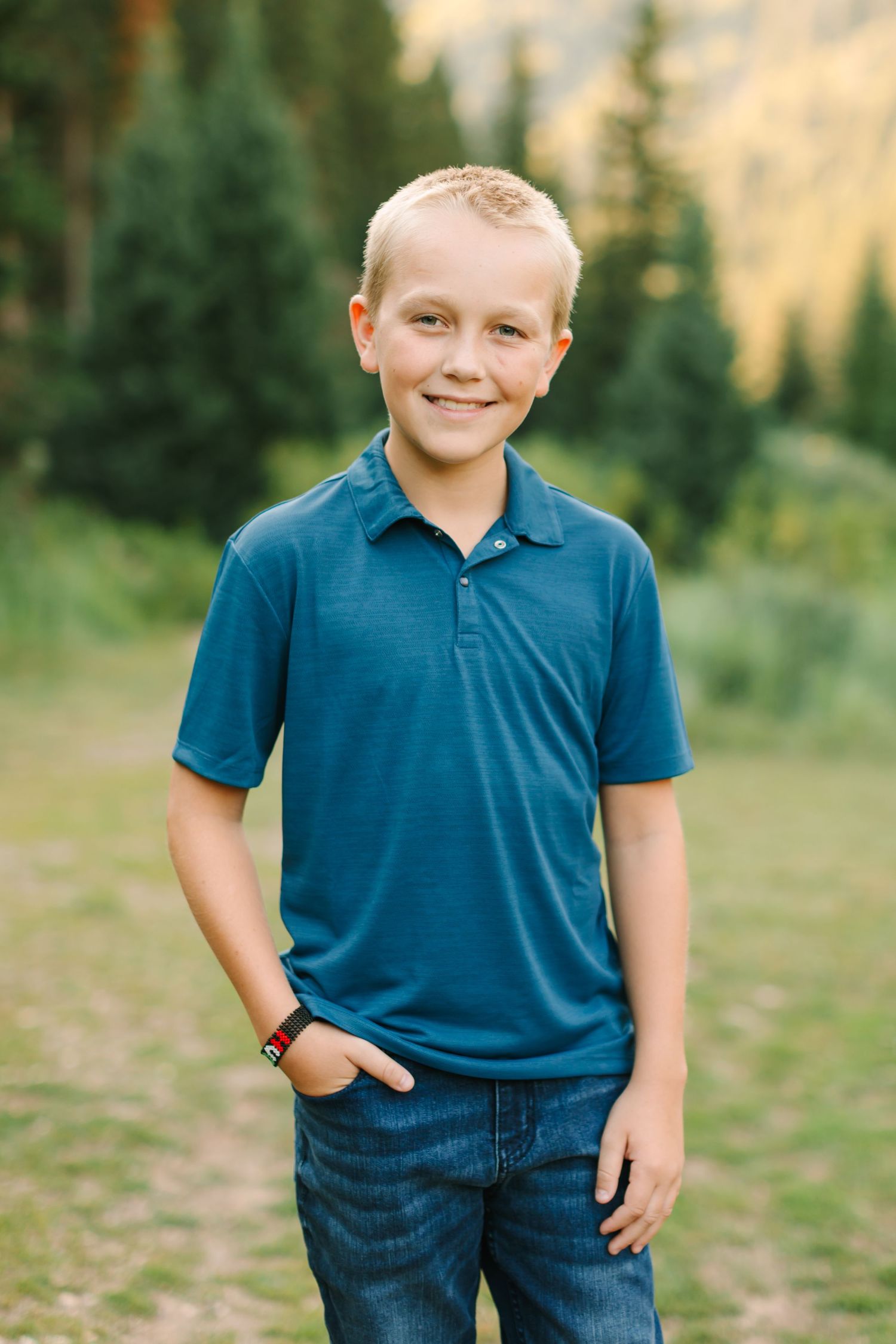 Smiling blonde boy in blue polo shirt and jeans poses outdoors with pine trees and mountains in background.