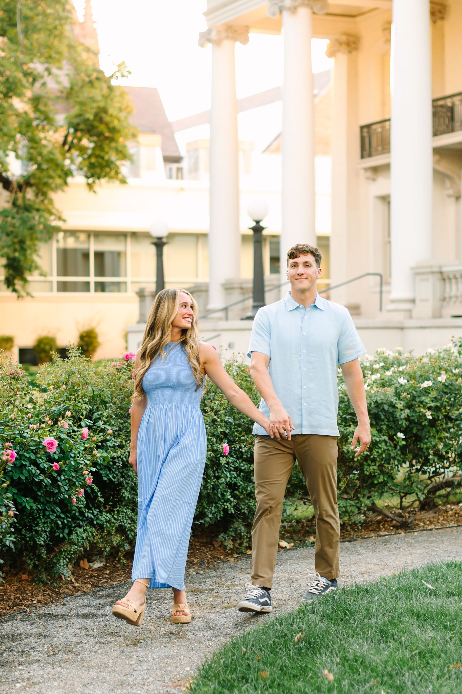 A young couple in coordinating blue outfits holds hands while walking through a garden path near a classical building.