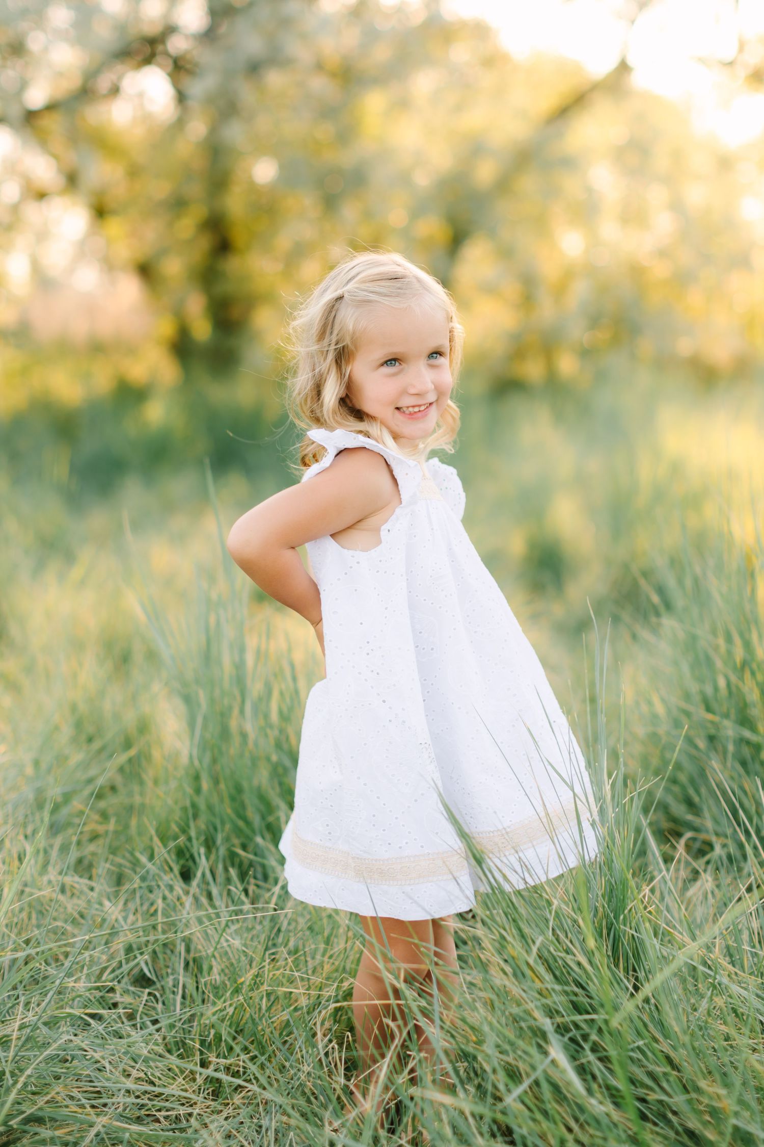 A smiling blonde girl in a white dress stands in tall green grass during golden hour.