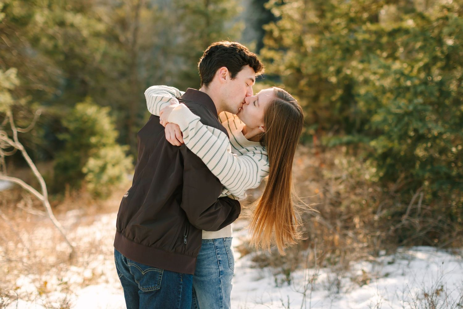 A couple embraces while standing in a snowy winter forest setting with golden sunlight filtering through the trees.