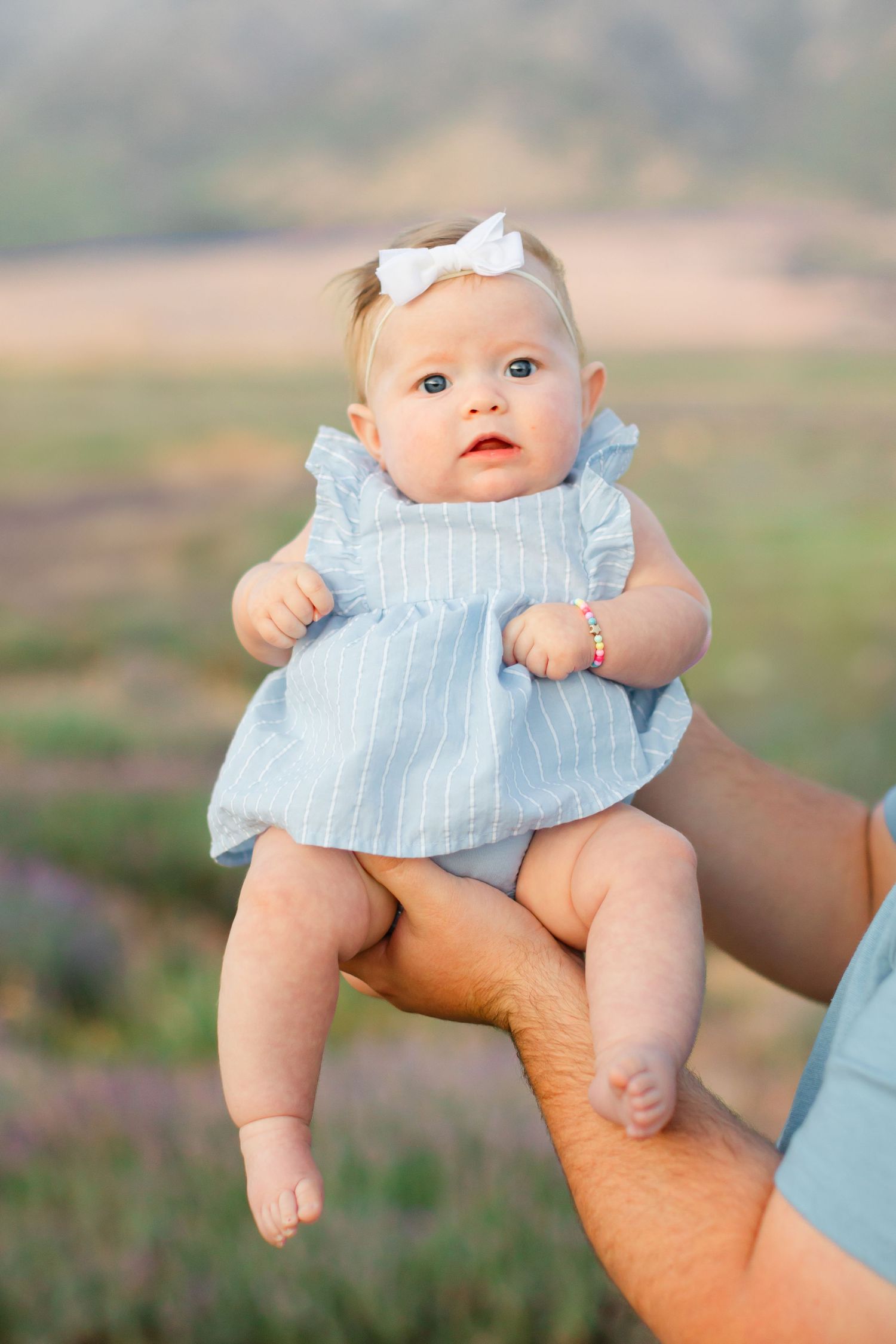 Baby wearing a light blue dress and white bow being held up outdoors against a blurred natural background.