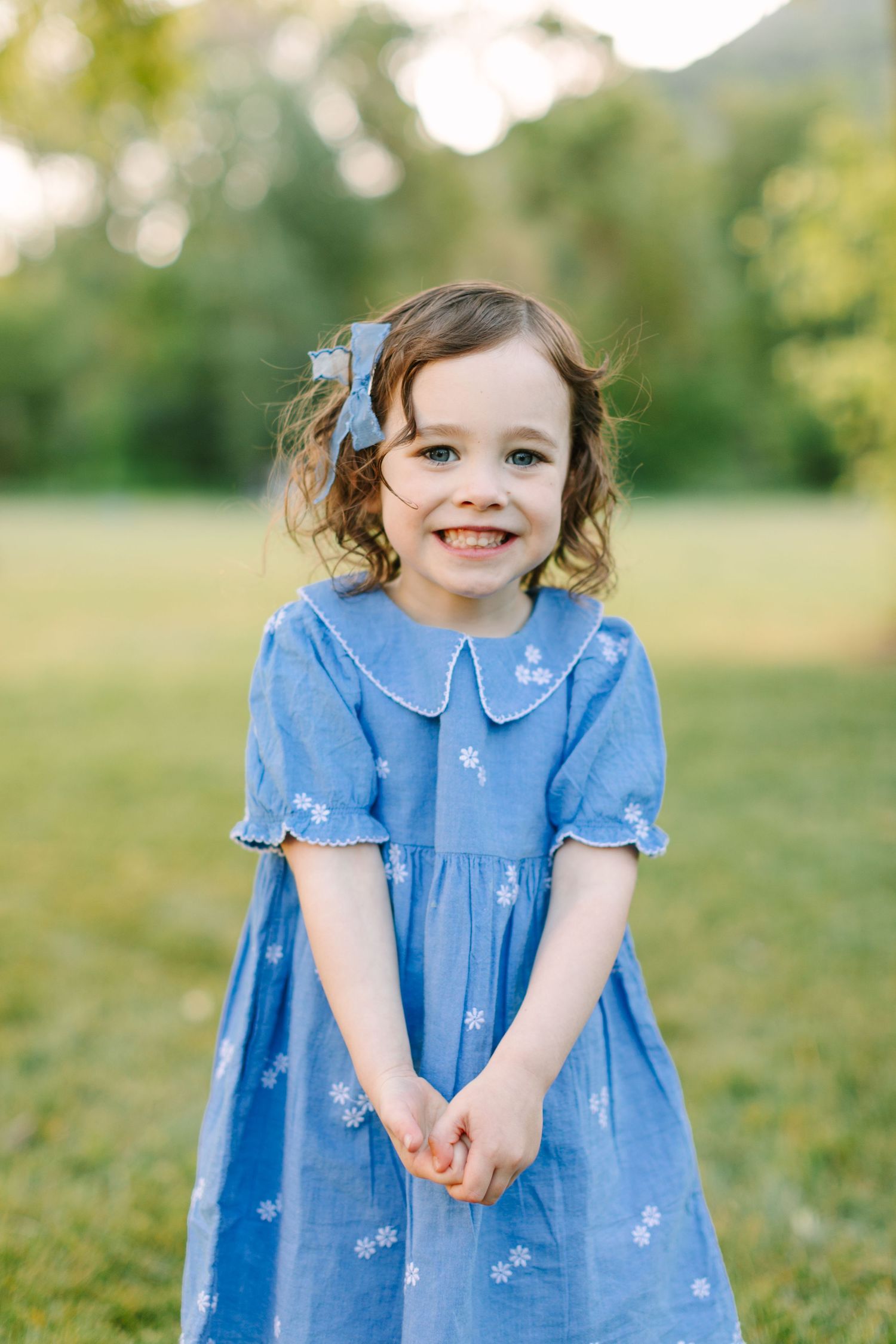 A young child in a blue dress with a matching hair bow stands smiling in a sunny outdoor setting.
