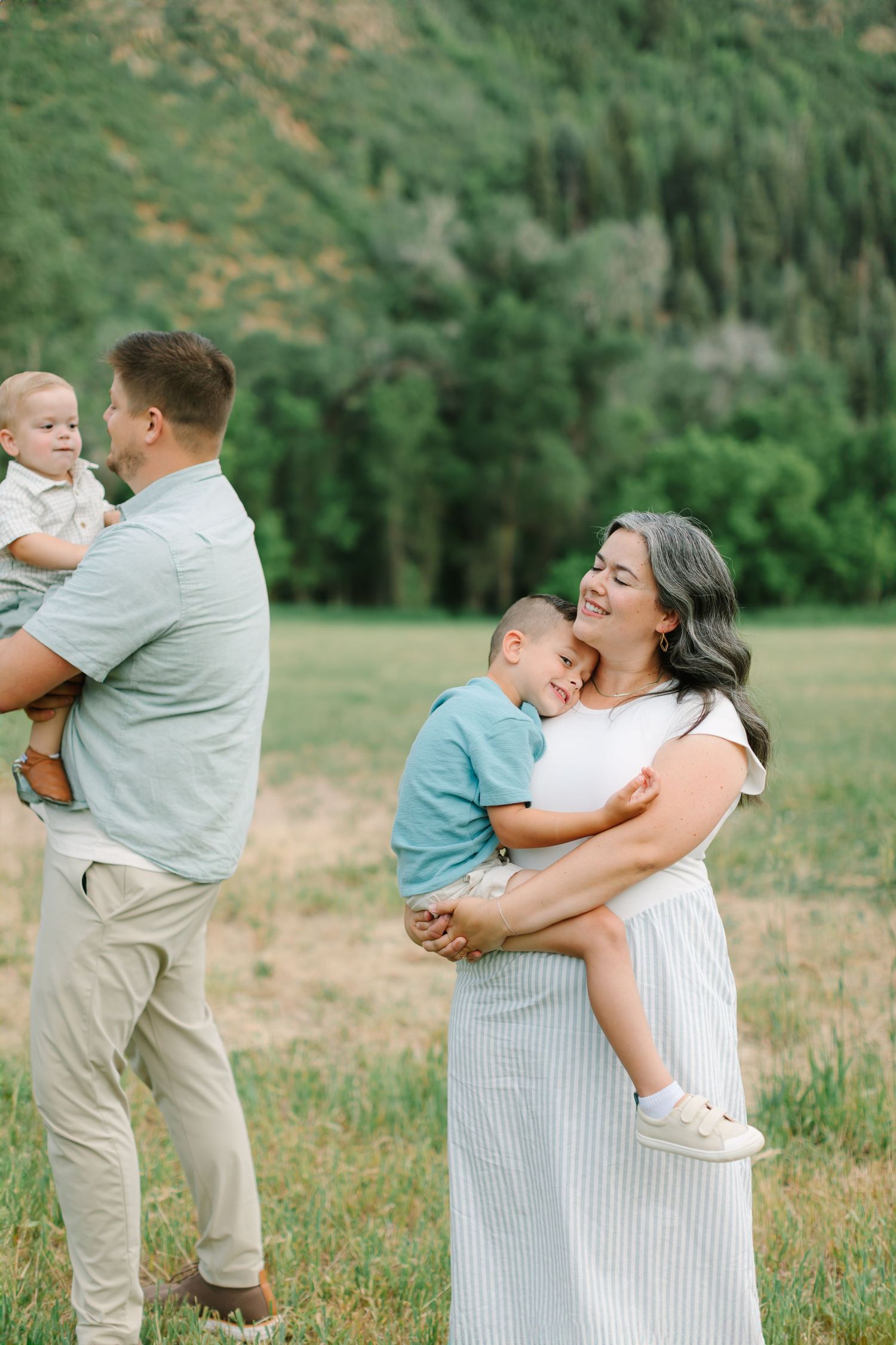 A family shares joyful embraces and tender moments together in a sunlit meadow with green foliage in the background.