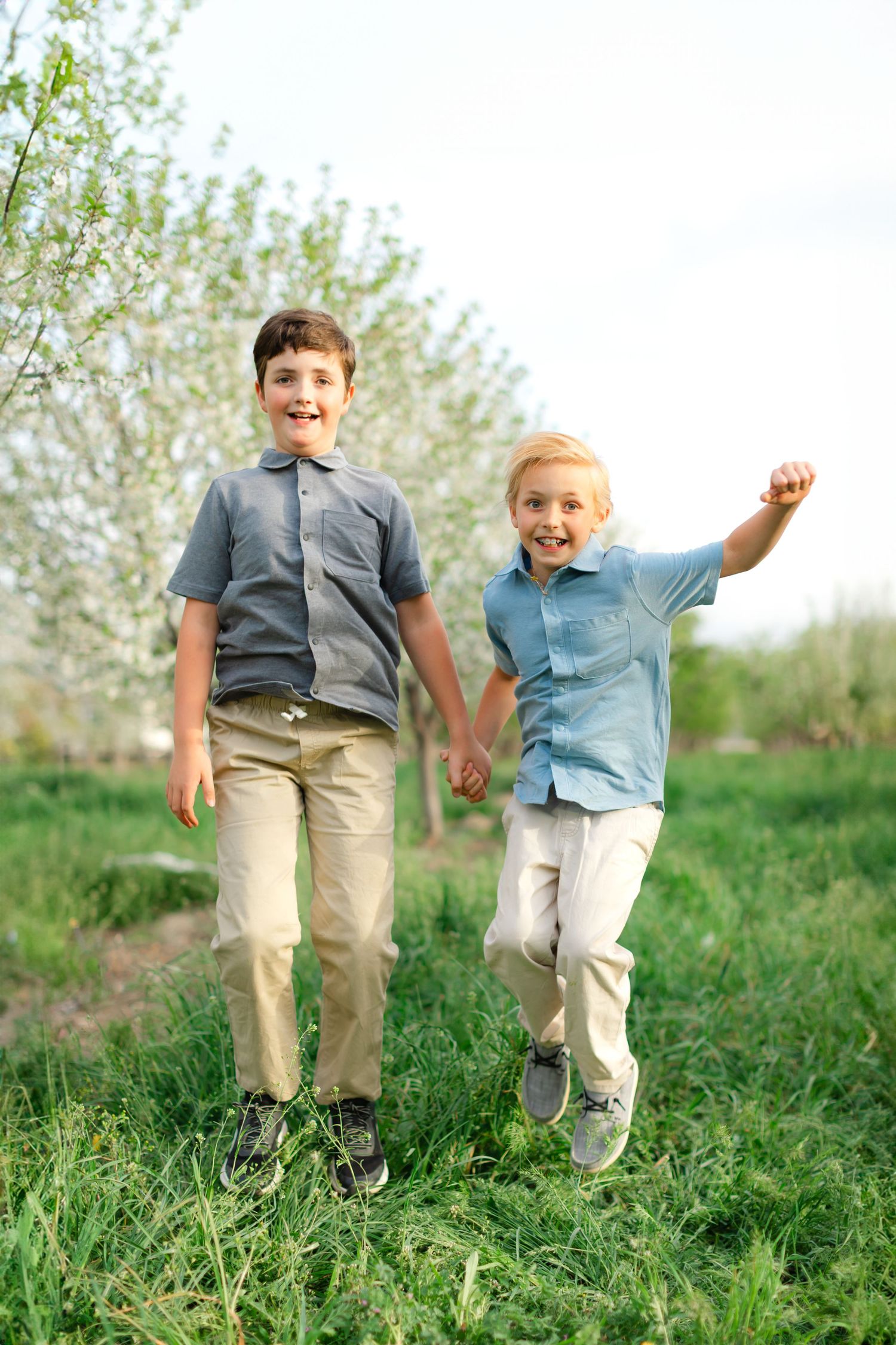Two children play and jump together in a grassy field on a sunny spring day with blooming trees in the background.