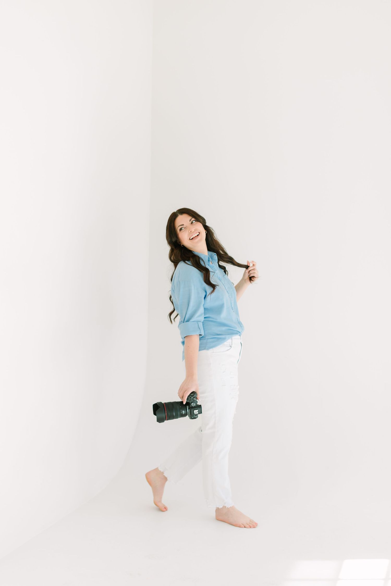Woman in light blue shirt and white pants poses playfully against white backdrop in lifestyle photoshoot series.