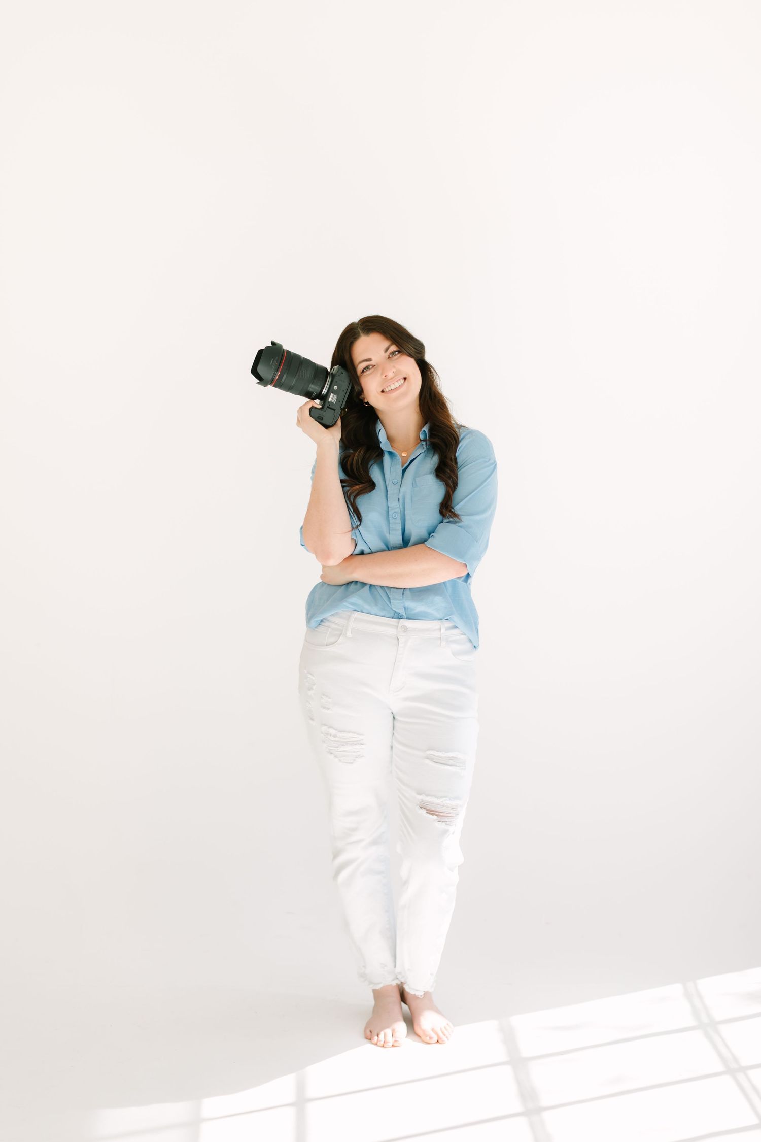 Professional photographer wearing denim shirt and white jeans holds camera while standing against white wall.