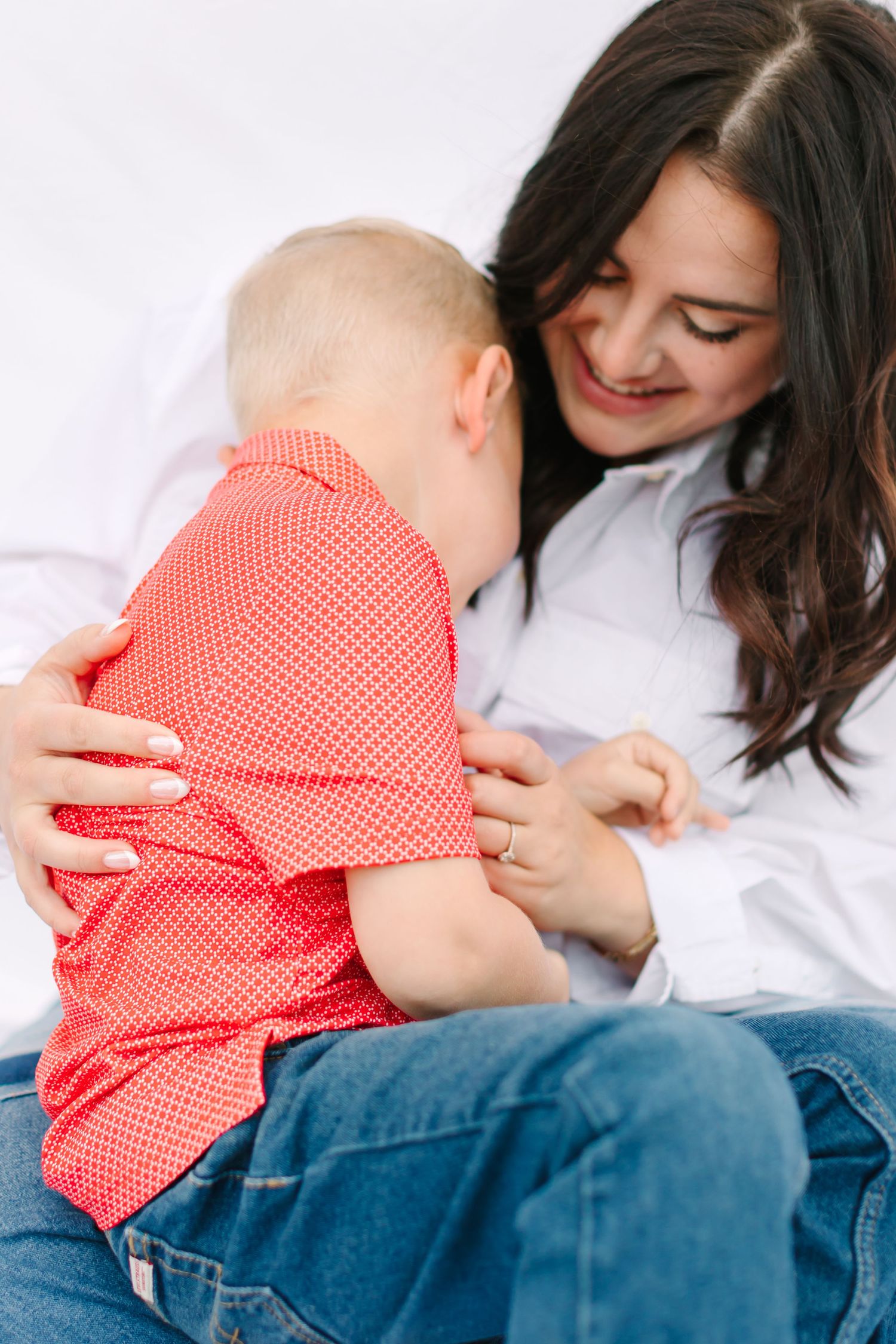 A smiling dark-haired mother in white shirt and jeans holds her baby boy in a red shirt on her lap.