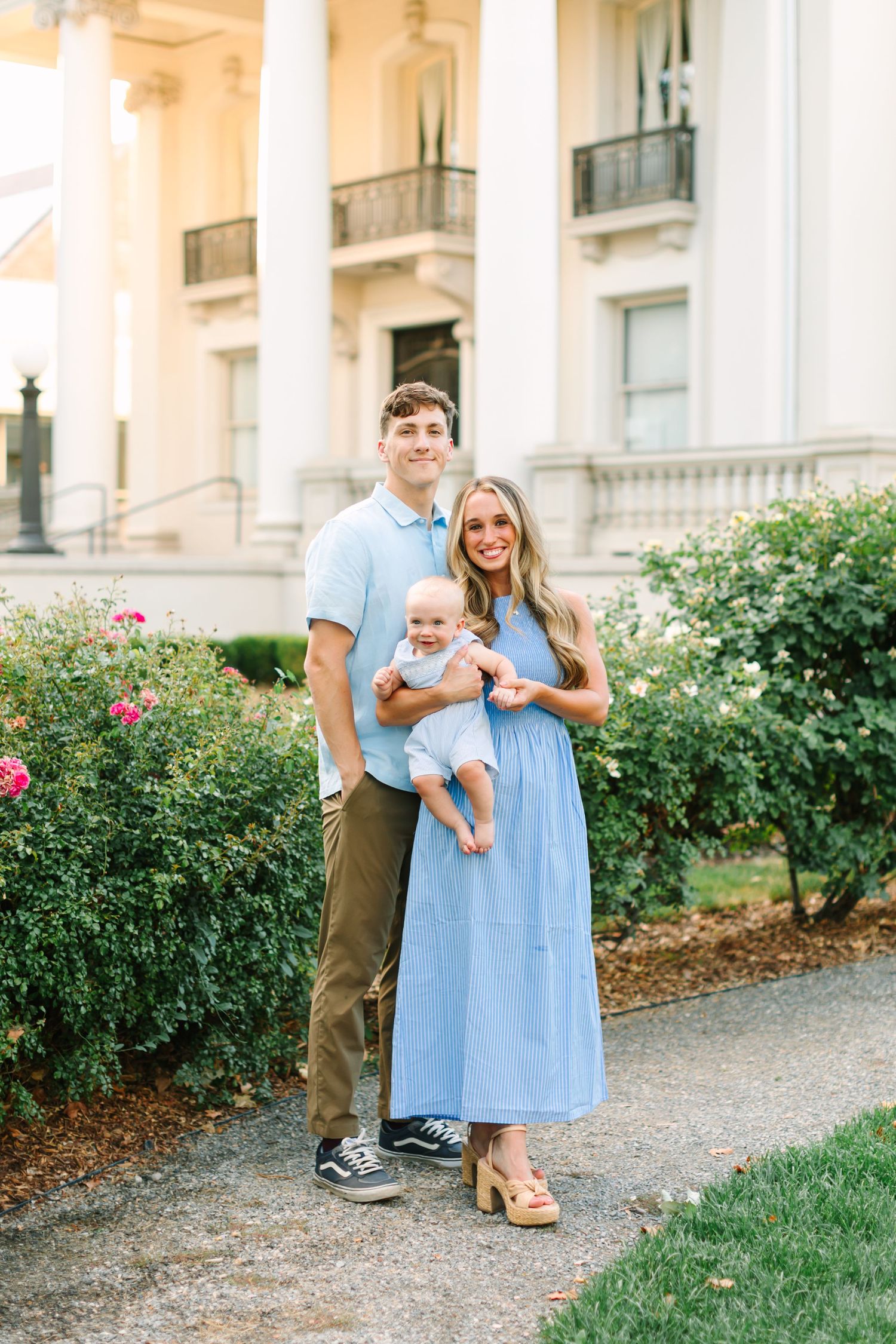Young family in coordinating blue outfits poses with baby outside elegant white columned mansion.