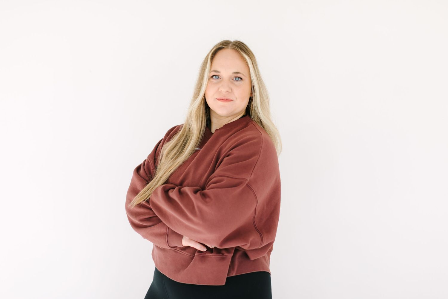 Person with long blonde hair wearing a rust-colored sweater stands with arms crossed against white background.