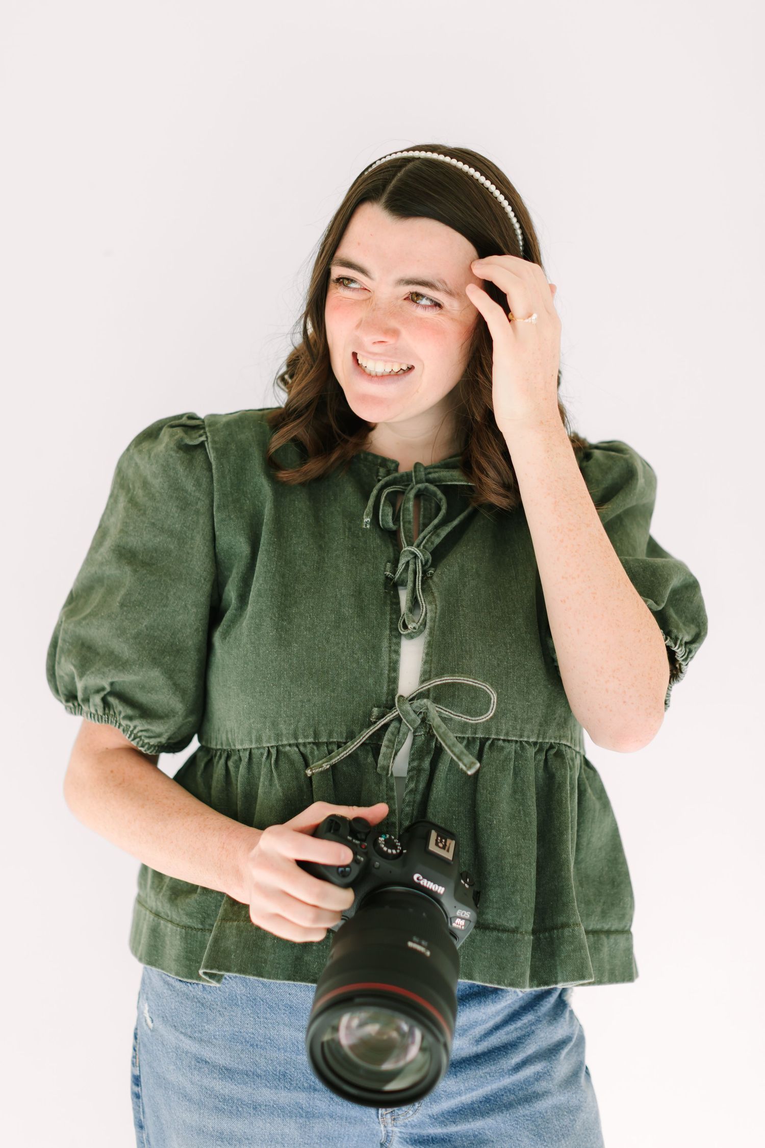 A photographer in a green top and jeans poses playfully with a DSLR camera against a white background.