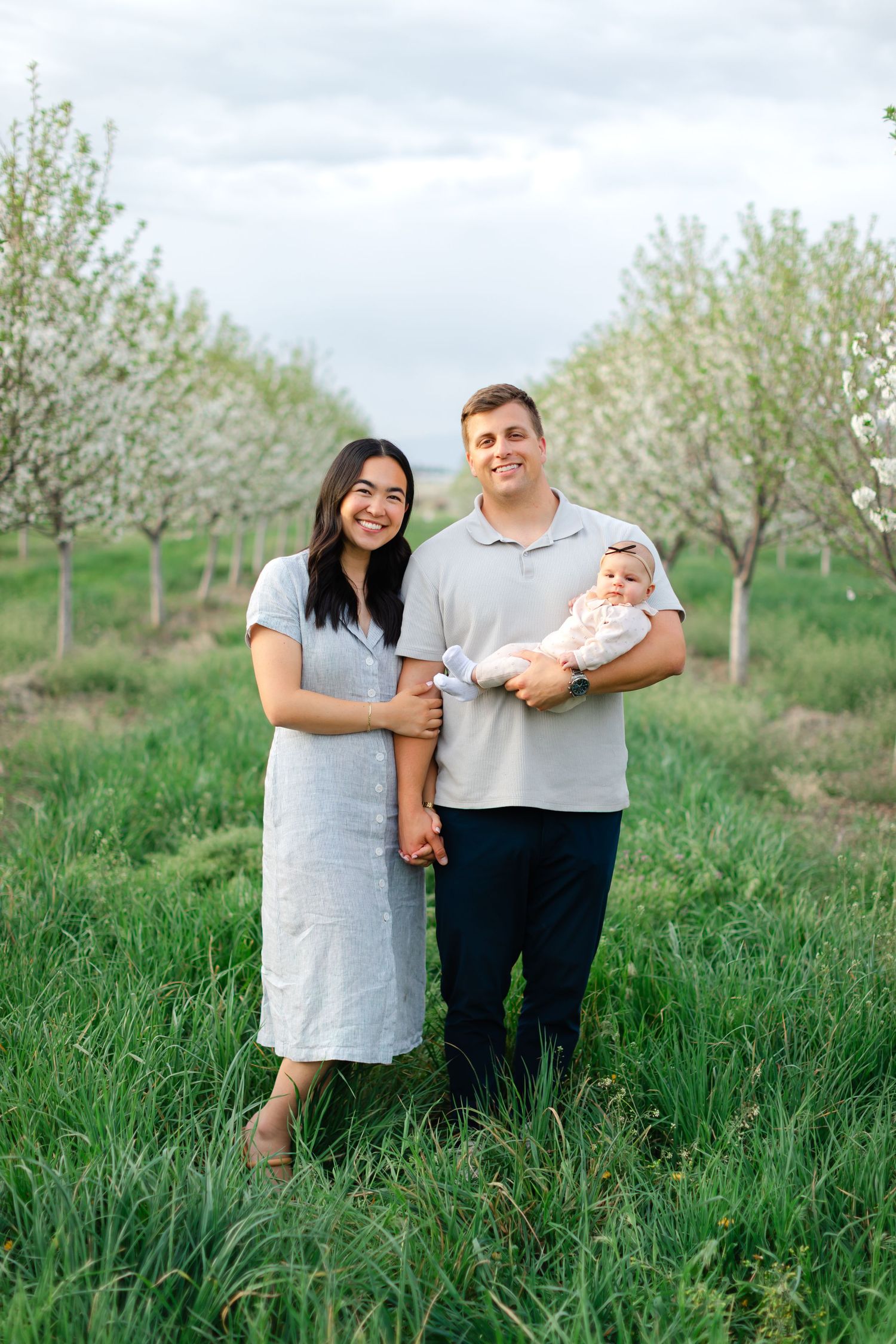 A couple stands with their baby in a blooming orchard surrounded by lush green grass and white flowering trees.