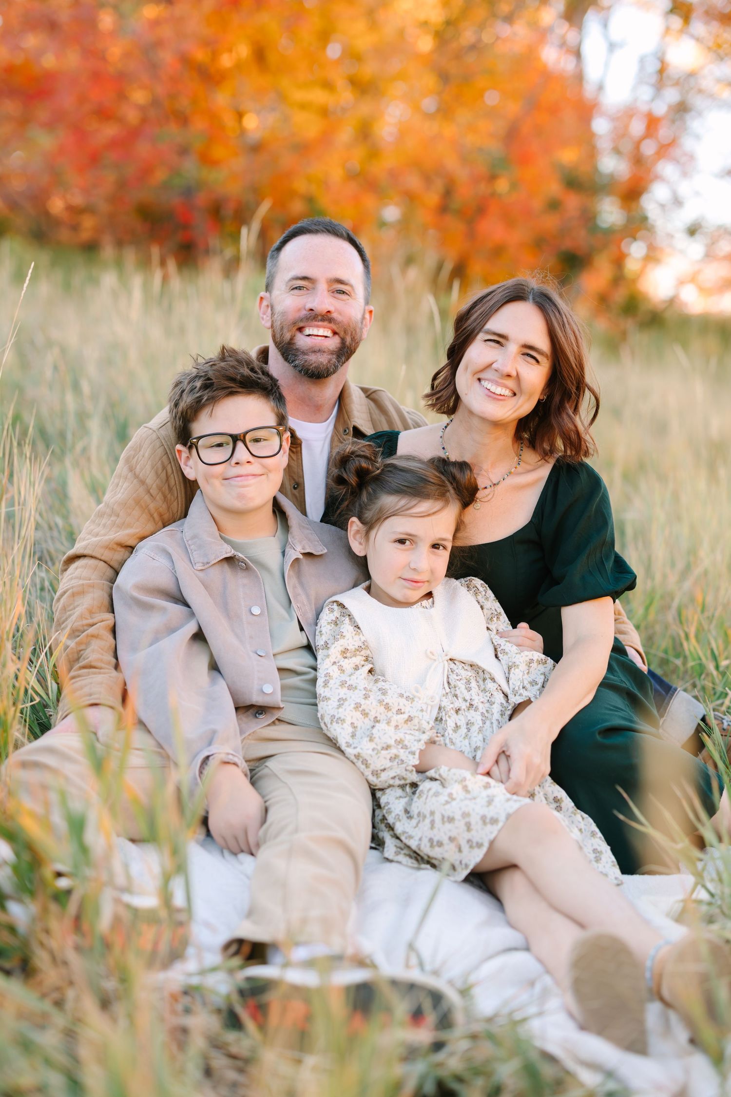 Family relaxes together in a golden autumn field with warm fall foliage in the background during sunset.