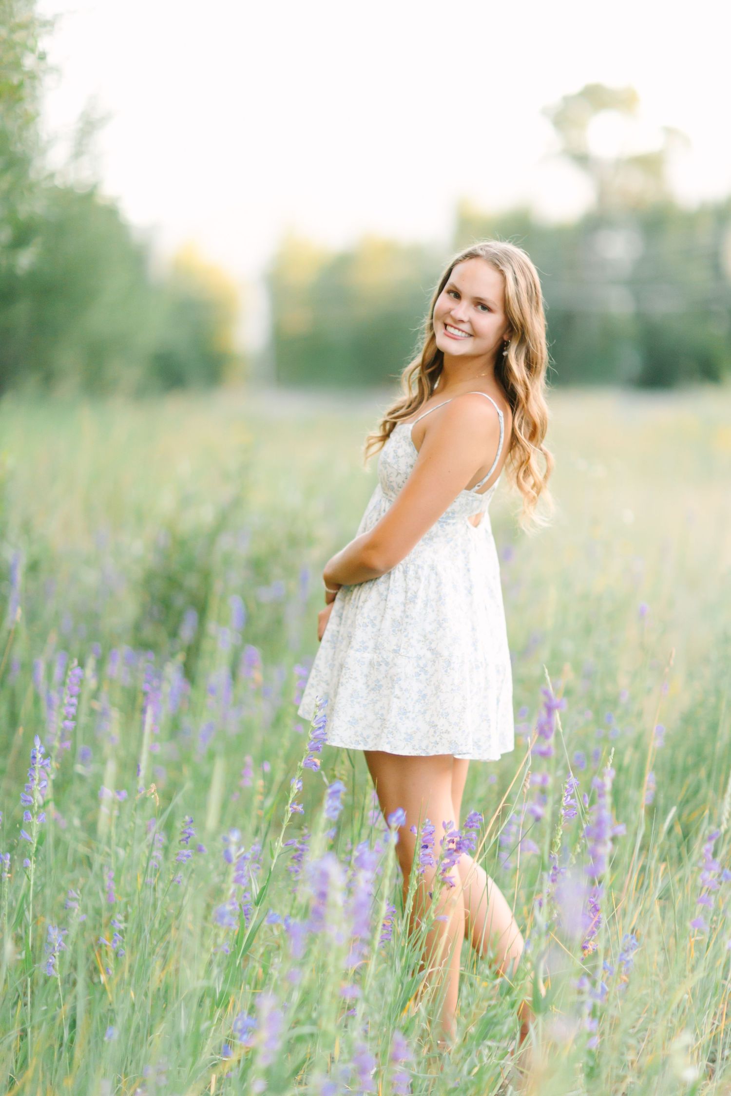 A person in a white sundress poses in a field of purple wildflowers during golden hour.