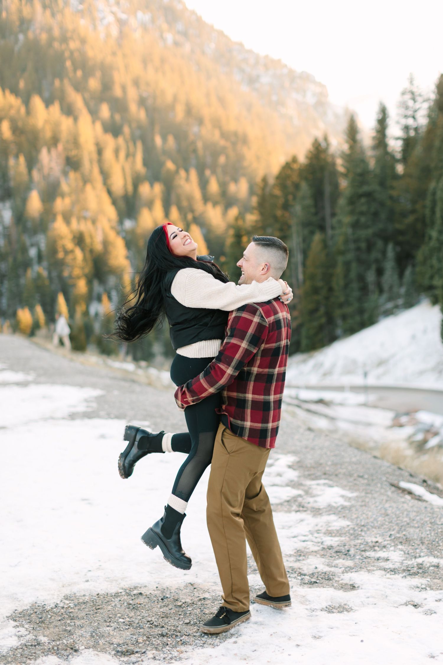 Playful couple in winter attire spins together on a snowy mountain path with evergreen trees and golden hills behind them.