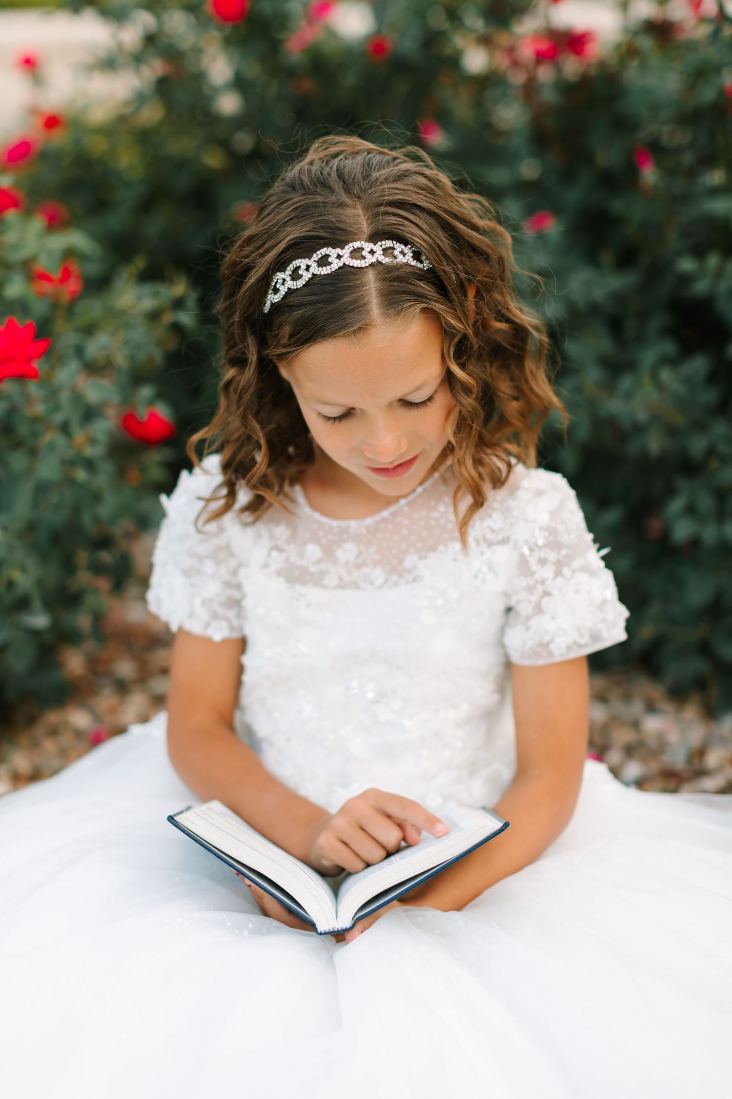 A young reader in a white lace dress and sparkly headband sits reading a book against a backdrop of red roses.