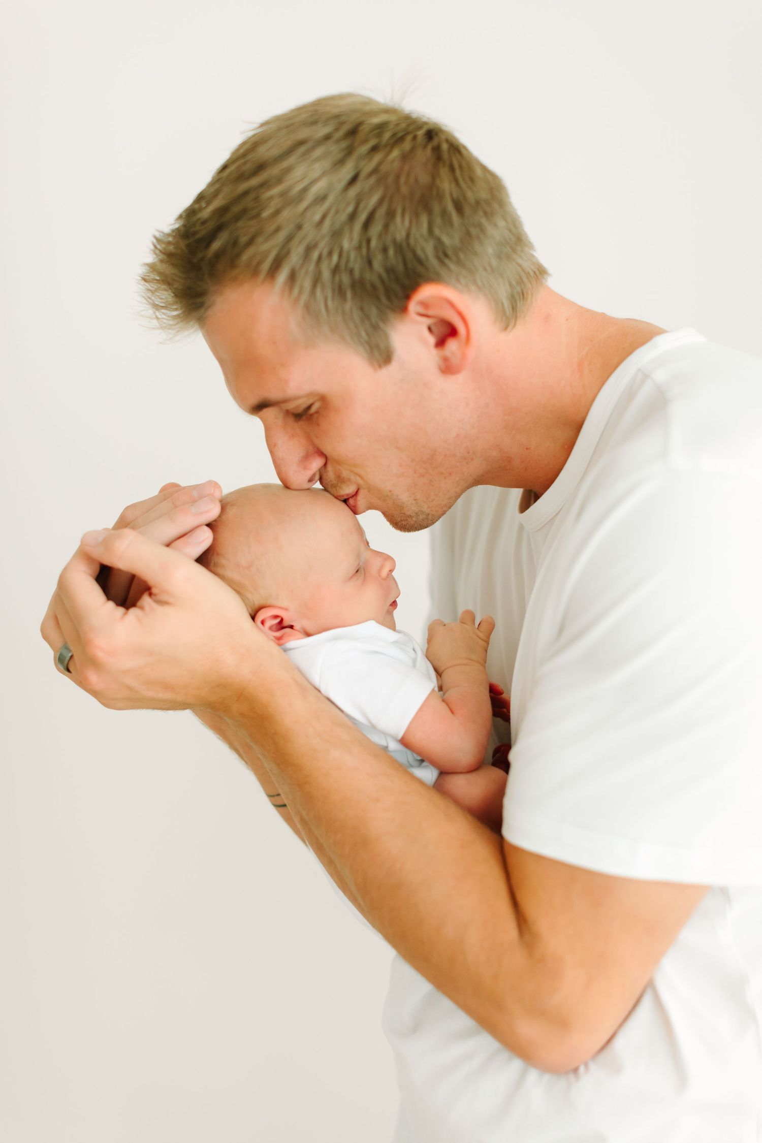 Parent in white shirt tenderly kisses newborn baby in intimate portrait series.