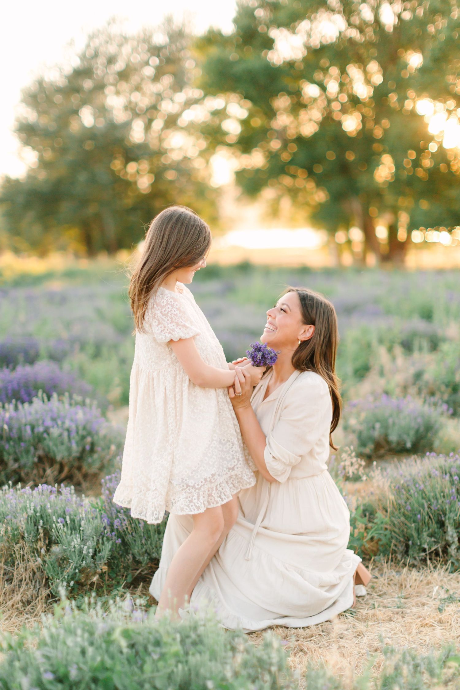 Two people in white dresses sit together in a blooming lavender field at golden hour.