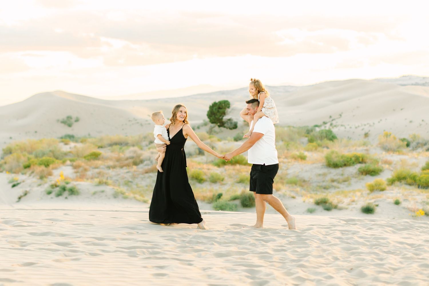A group of dancers in flowing black dresses and beige outfits perform gracefully on a sandy beach at sunset.