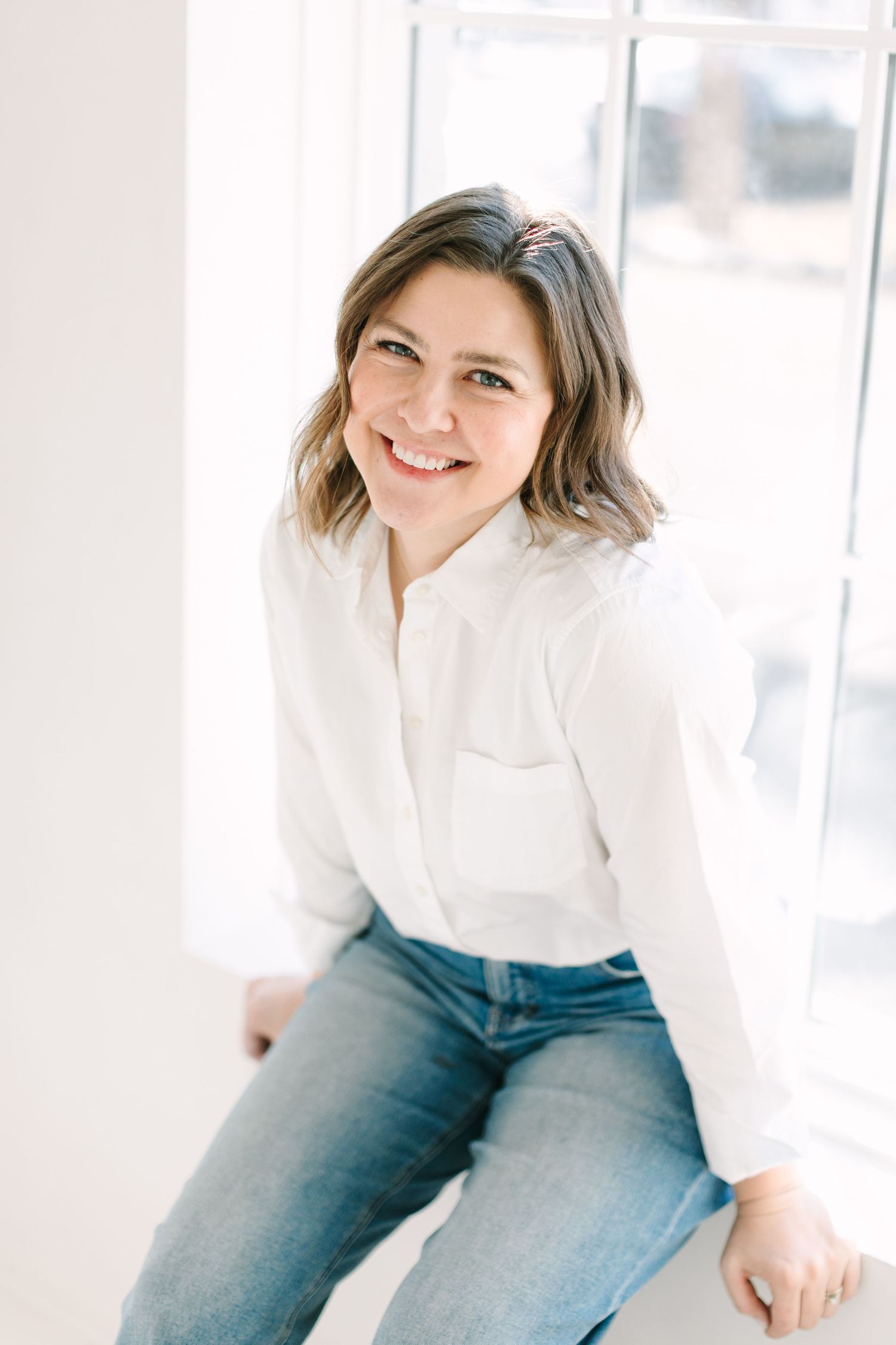 Professional lifestyle portrait of someone in a white button-down shirt and blue jeans sitting casually by a window.