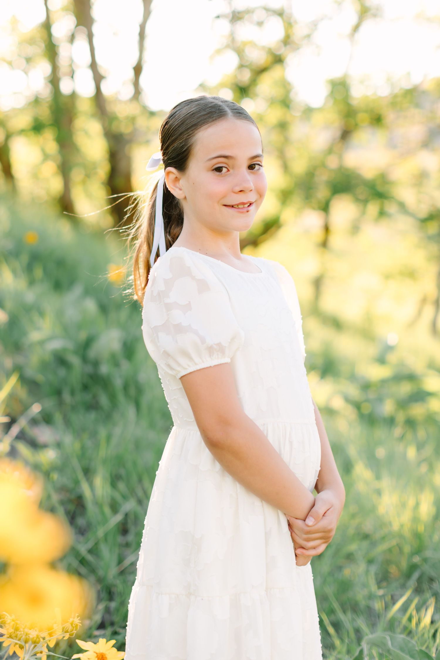 Young girl in white lace dress smiling outdoors in green meadow with yellow wildflowers and golden sunlight.