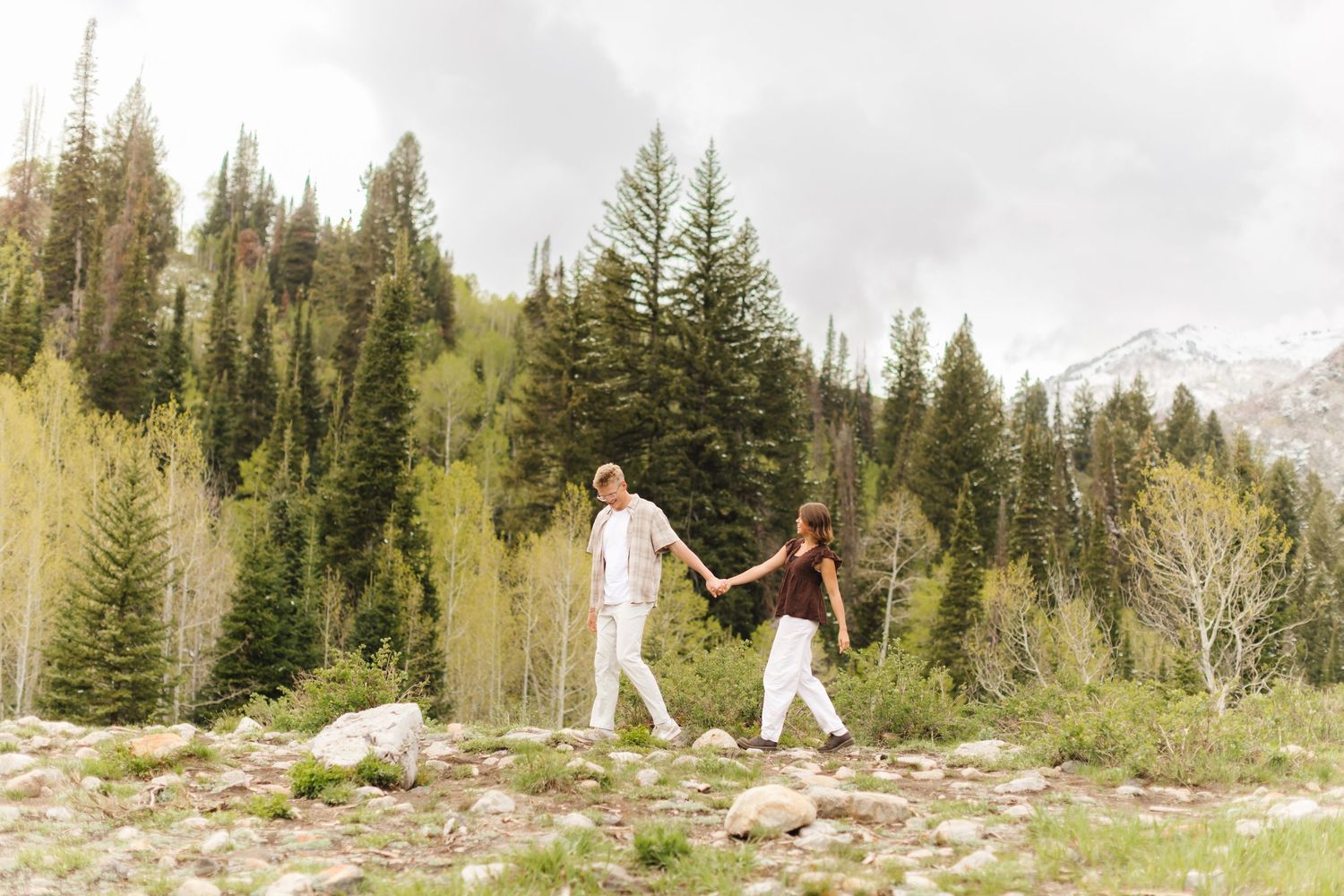 Couple holding hands walking through a rocky mountain meadow surrounded by evergreen and aspen trees in Salt Lake City, Utah.