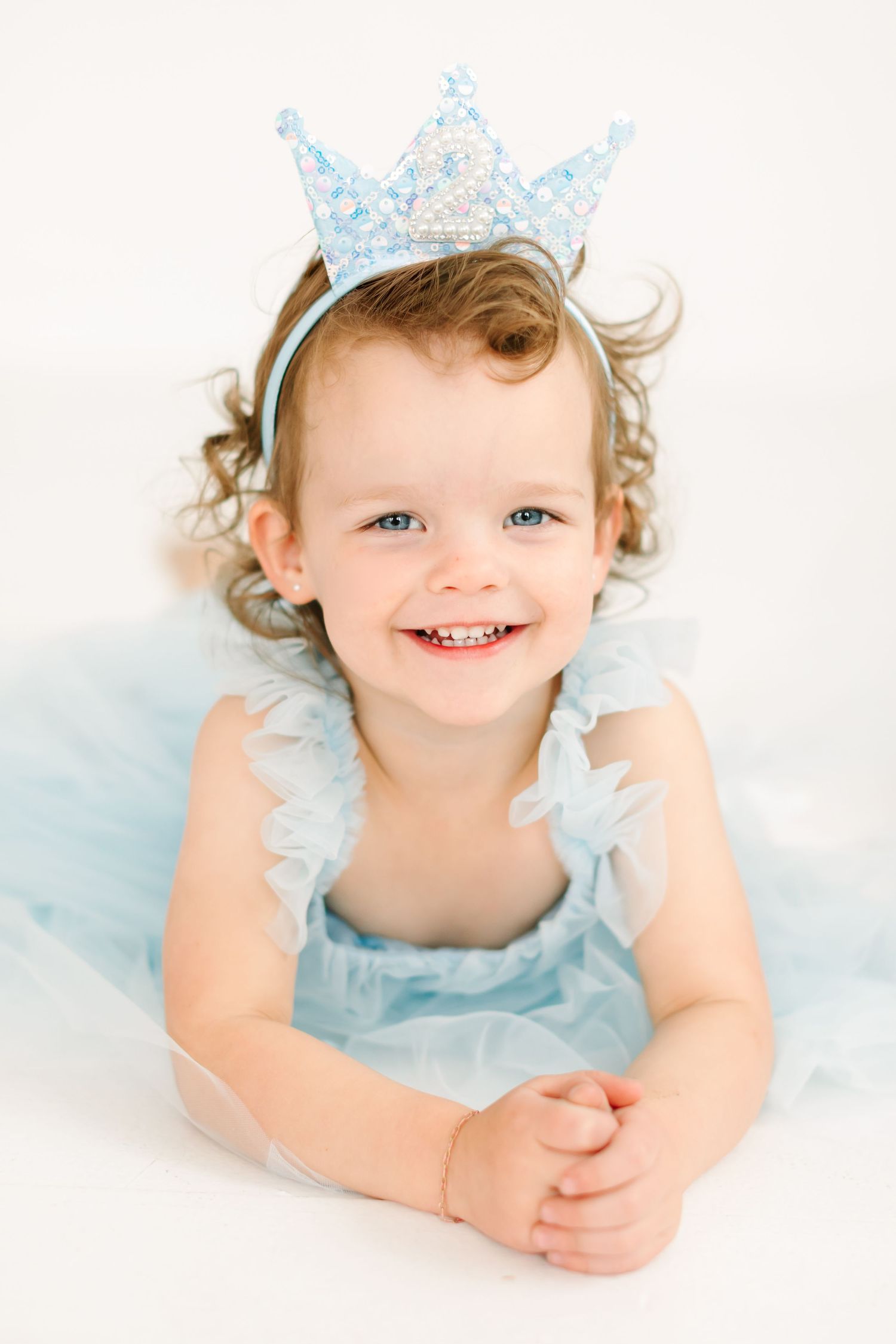 A smiling young girl wearing a blue dress and a crown-like headband, sitting on a white background.