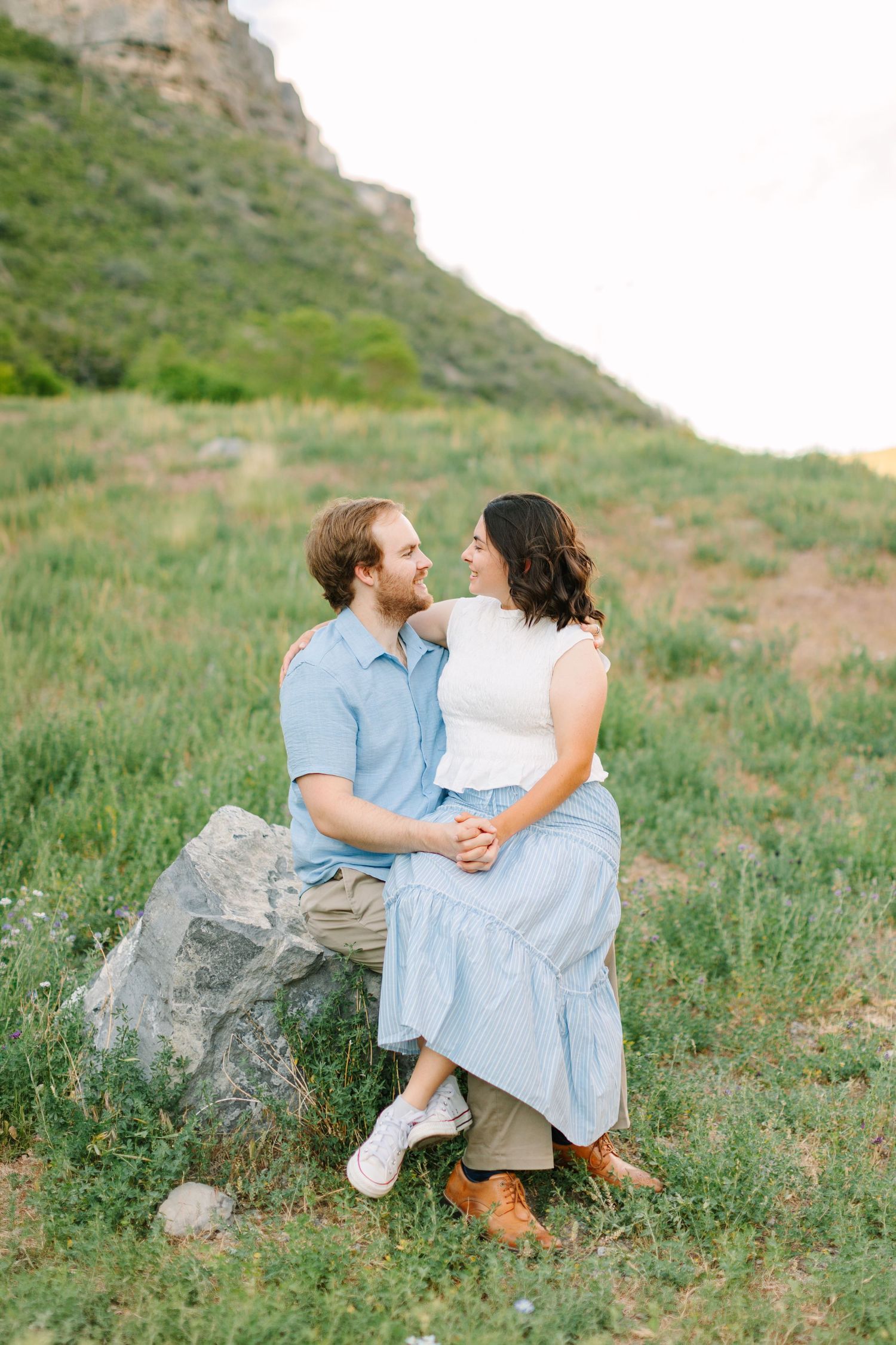 Couple sitting on a rock in a green meadow with mountains, smiling at each other in coordinating blue outfits.