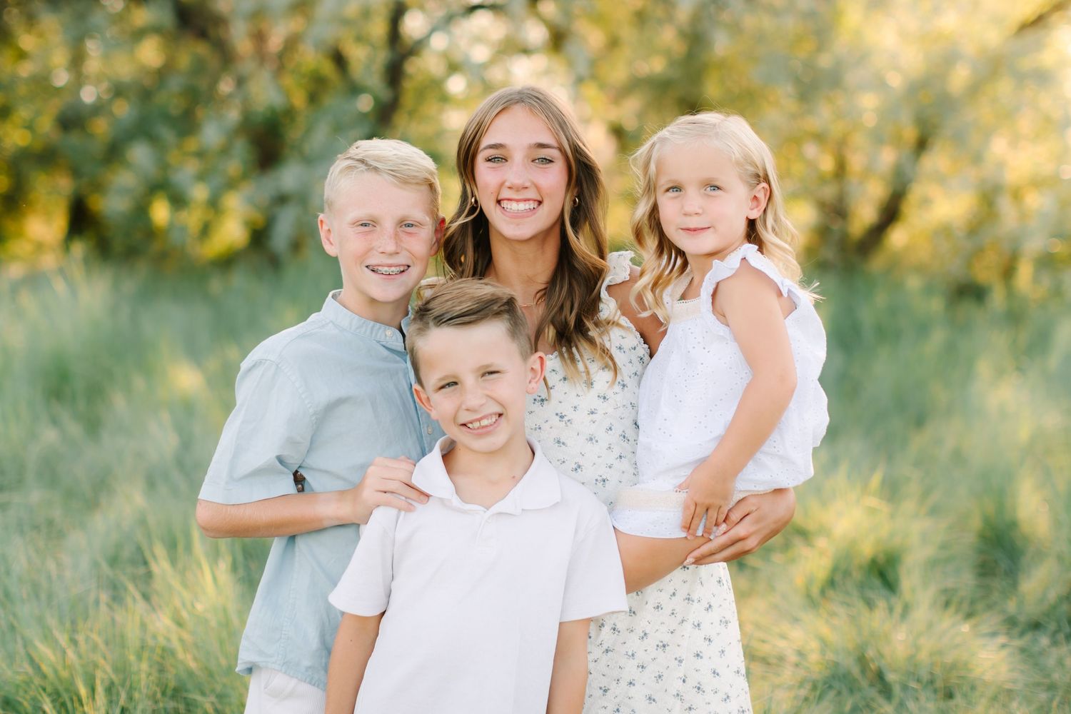 A smiling woman poses outdoors with three children in a sunny green field during a family portrait session.