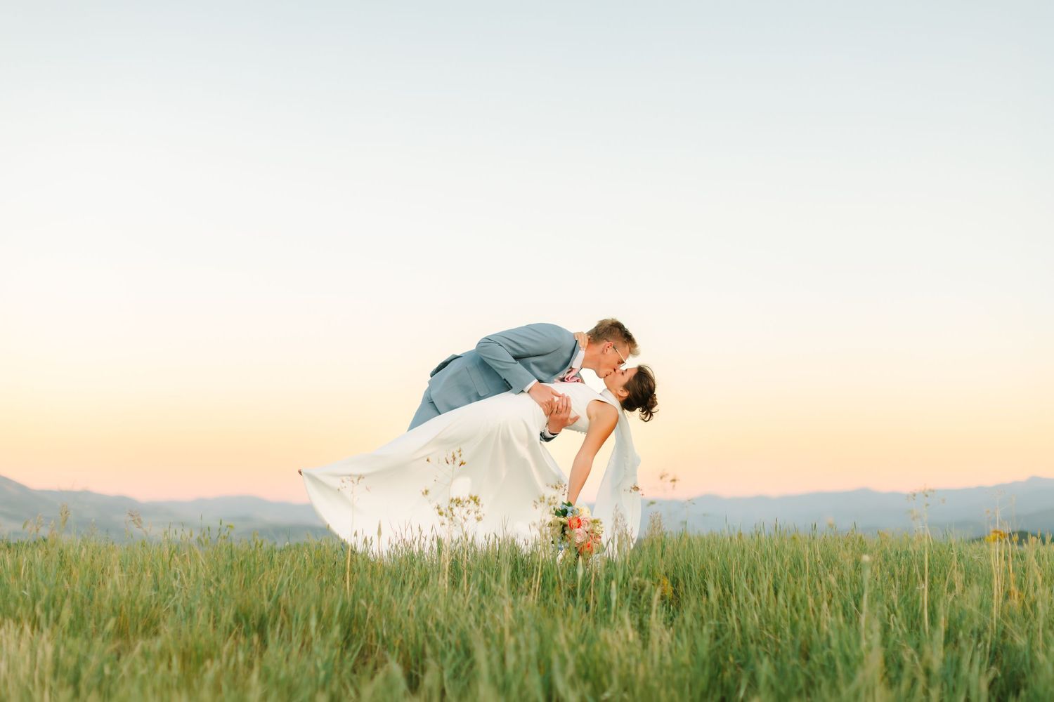Bride and groom share a romantic dip kiss in a green field at sunset with mountain views.