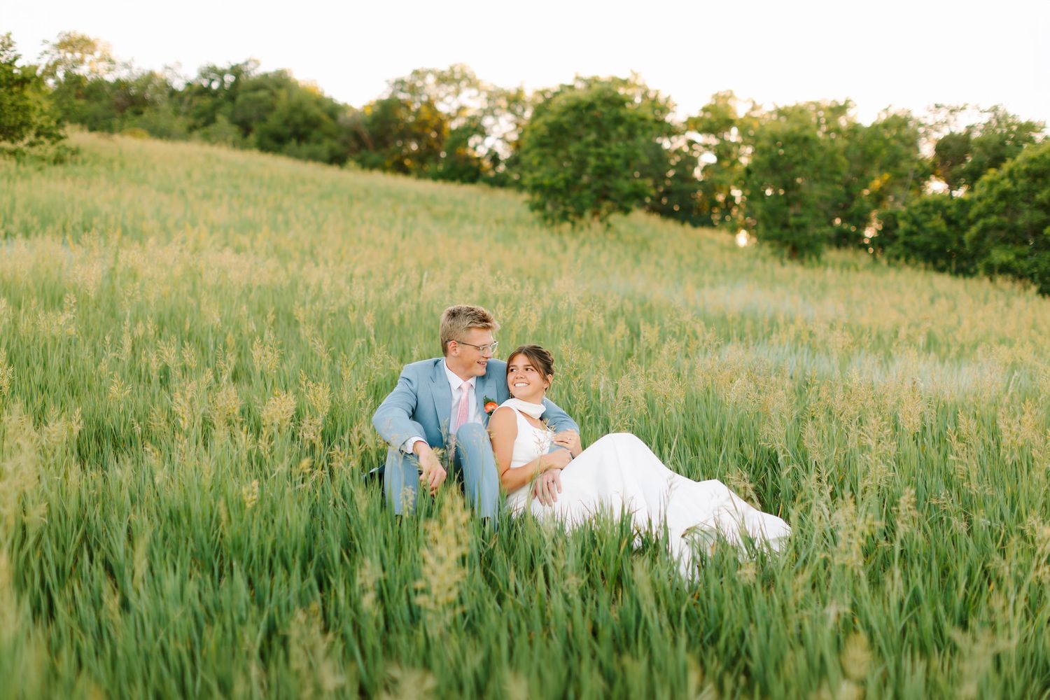 Bride and groom sitting together in a lush green field during golden hour at their outdoor wedding.