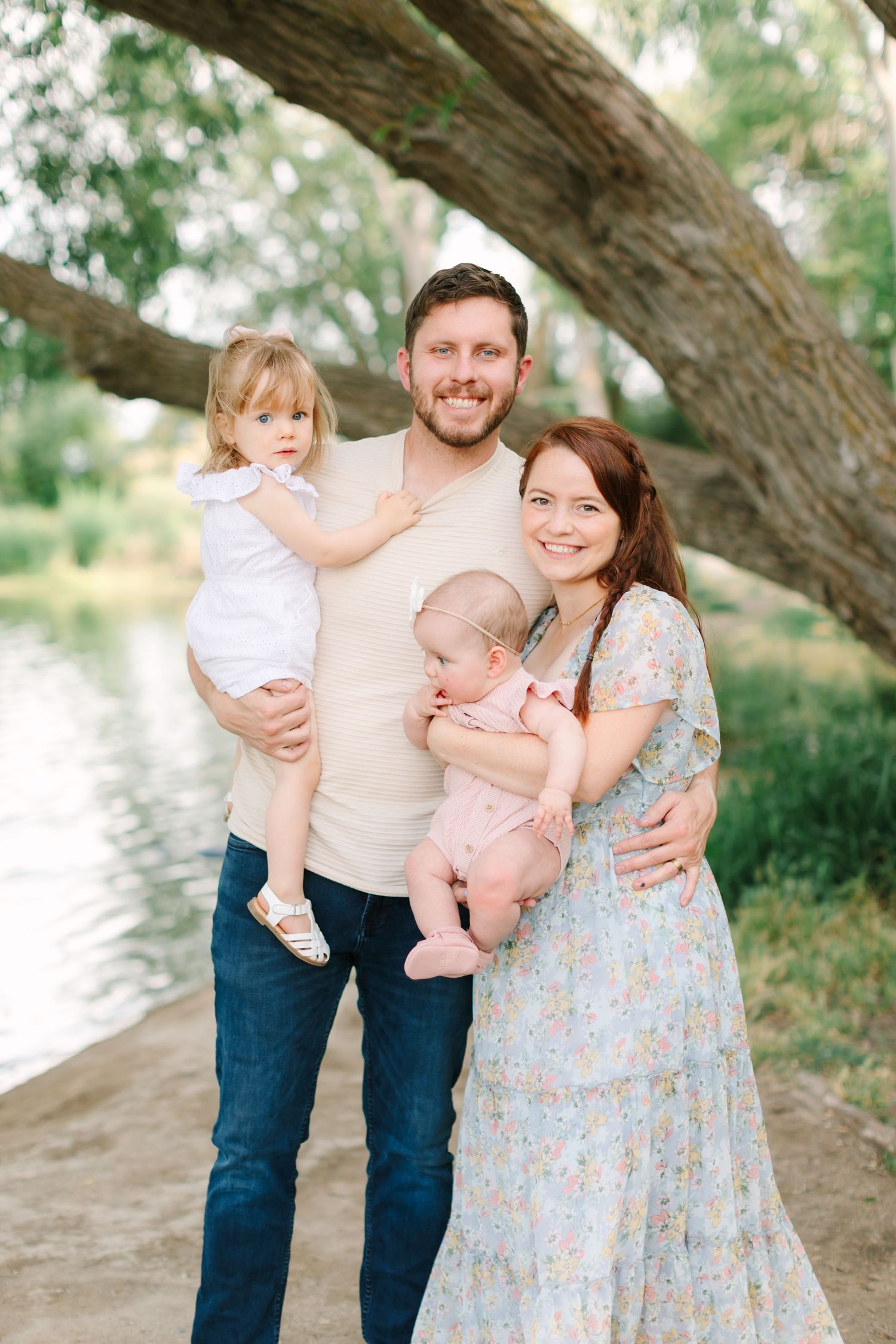 Happy family of four posing outdoors near a river under a large tree, parents holding two young children.