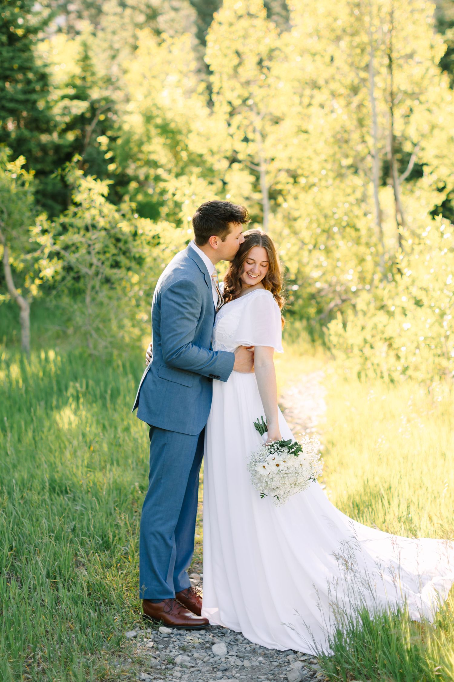 Bride and groom embrace in a sunlit forest, she holds a white bouquet in a flowing gown, he wears a blue suit.