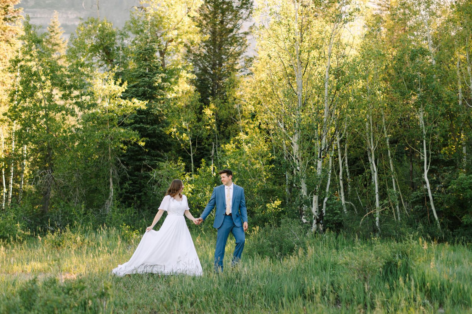 Bride and groom walk hand-in-hand through a lush green meadow surrounded by birch trees on their wedding day.